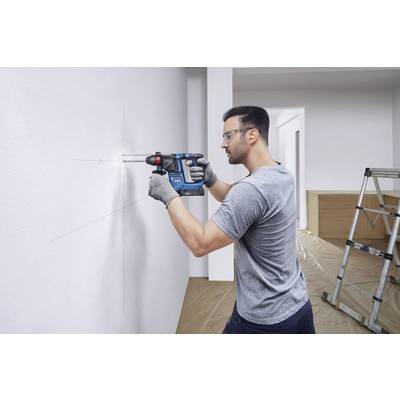 A man is drilling a hole in a white wall using a blue electric tool. A ladder is standing in the background, and the floor is covered.