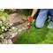 A person is kneeling on the lawn and trimming the grass edge along a stone path with a red garden shears. Flowers are blooming on the left.