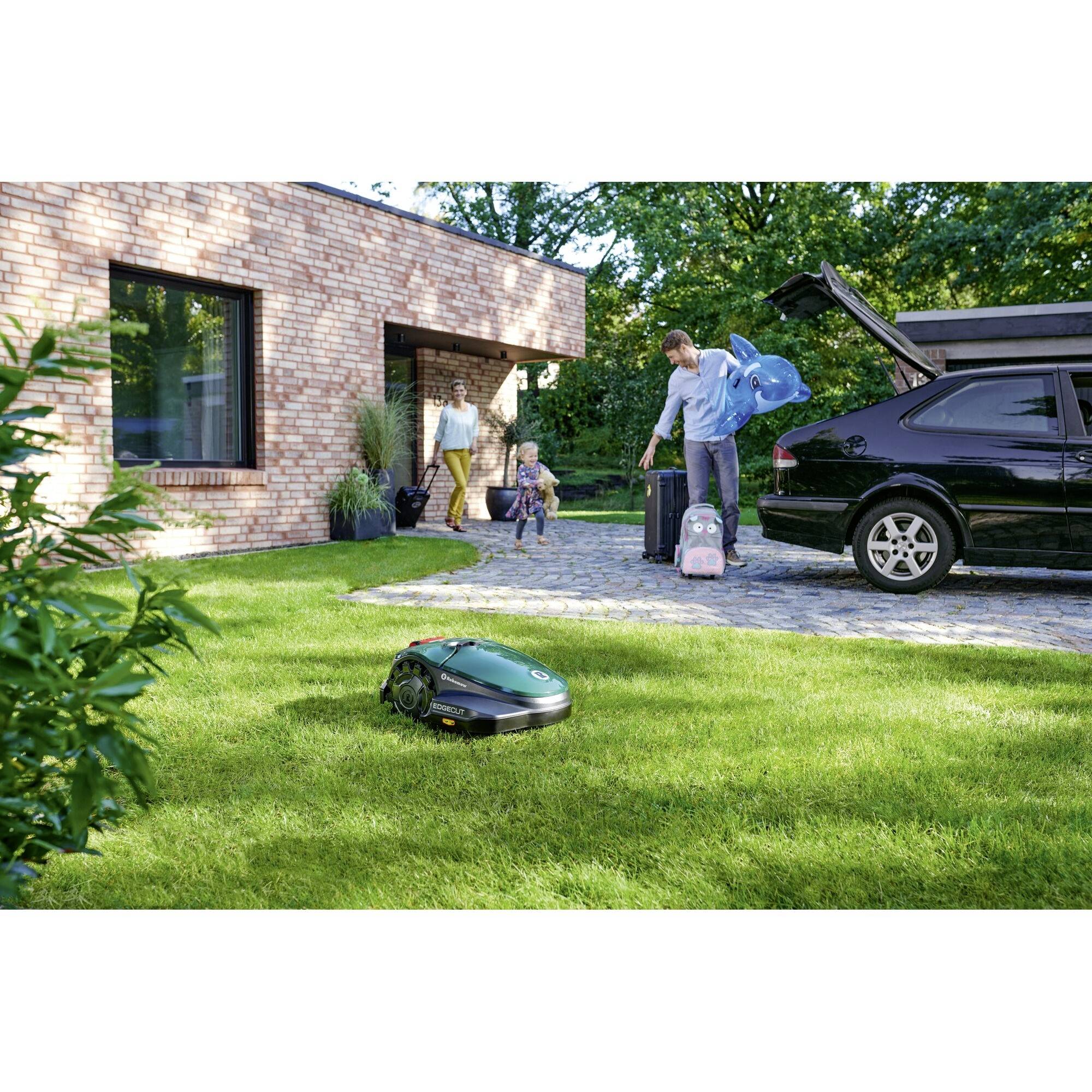A man is packing a suitcase into the car. His wife and child are sitting in the background on the grass. An automatic lawnmower is cutting the lawn. The family trip is being prepared.