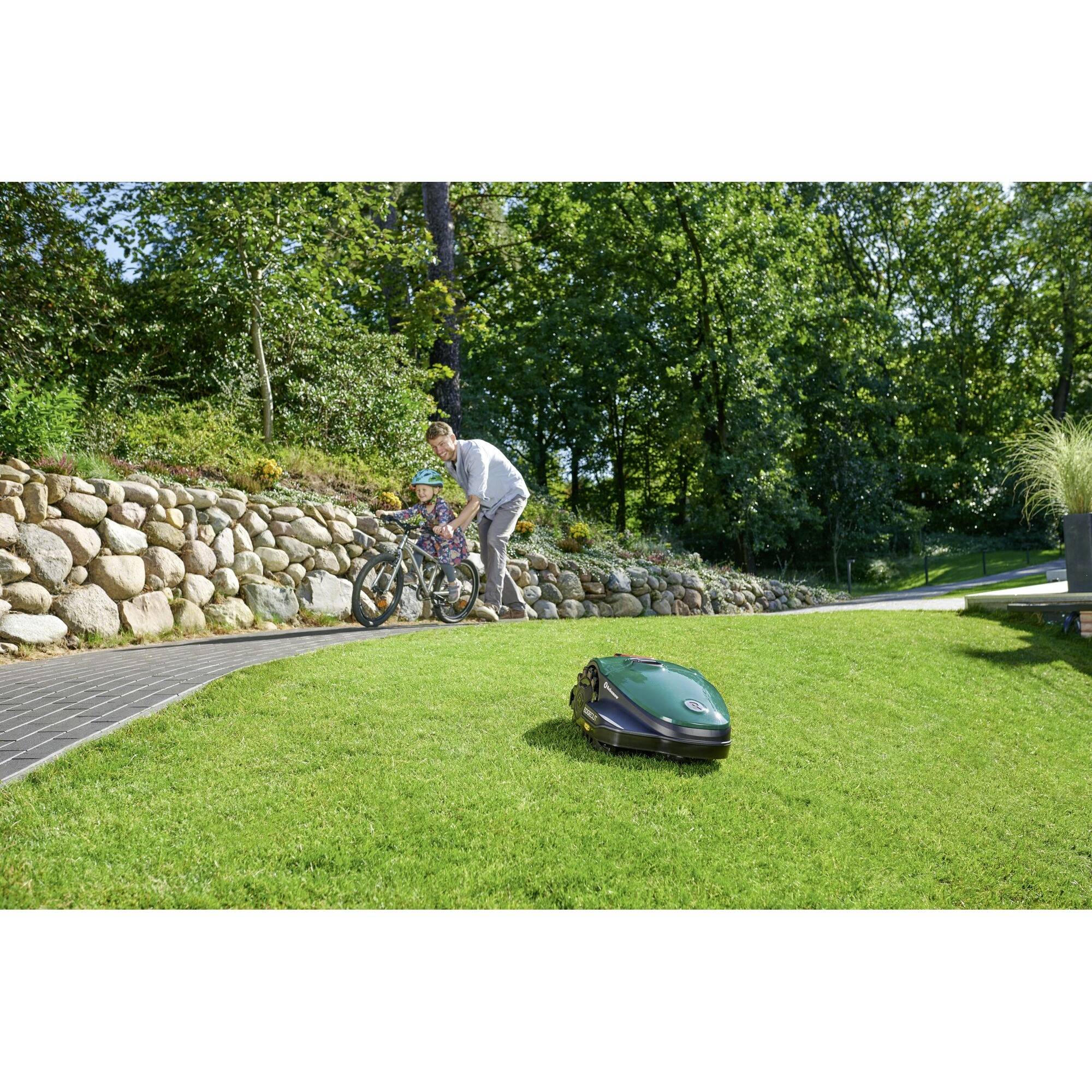 A robotic lawnmower is cutting grass in the foreground. In the background, a person is helping a child on a bicycle along a paved path.