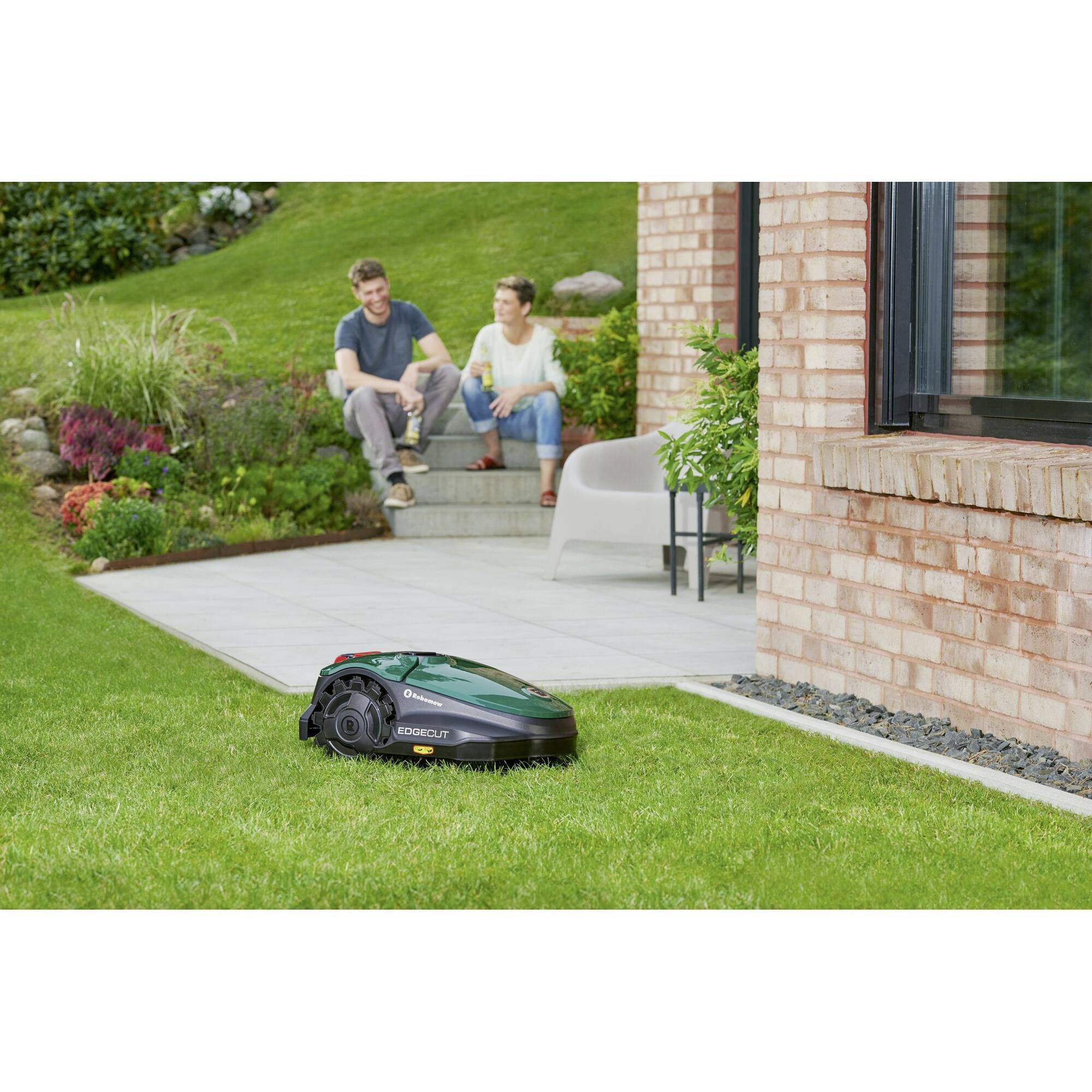 Robotic lawnmower mowing grass beside a brick building; two people sit on steps in the background, chatting.