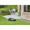 Robotic lawnmower mowing grass beside a brick building; two people sit on steps in the background, chatting.