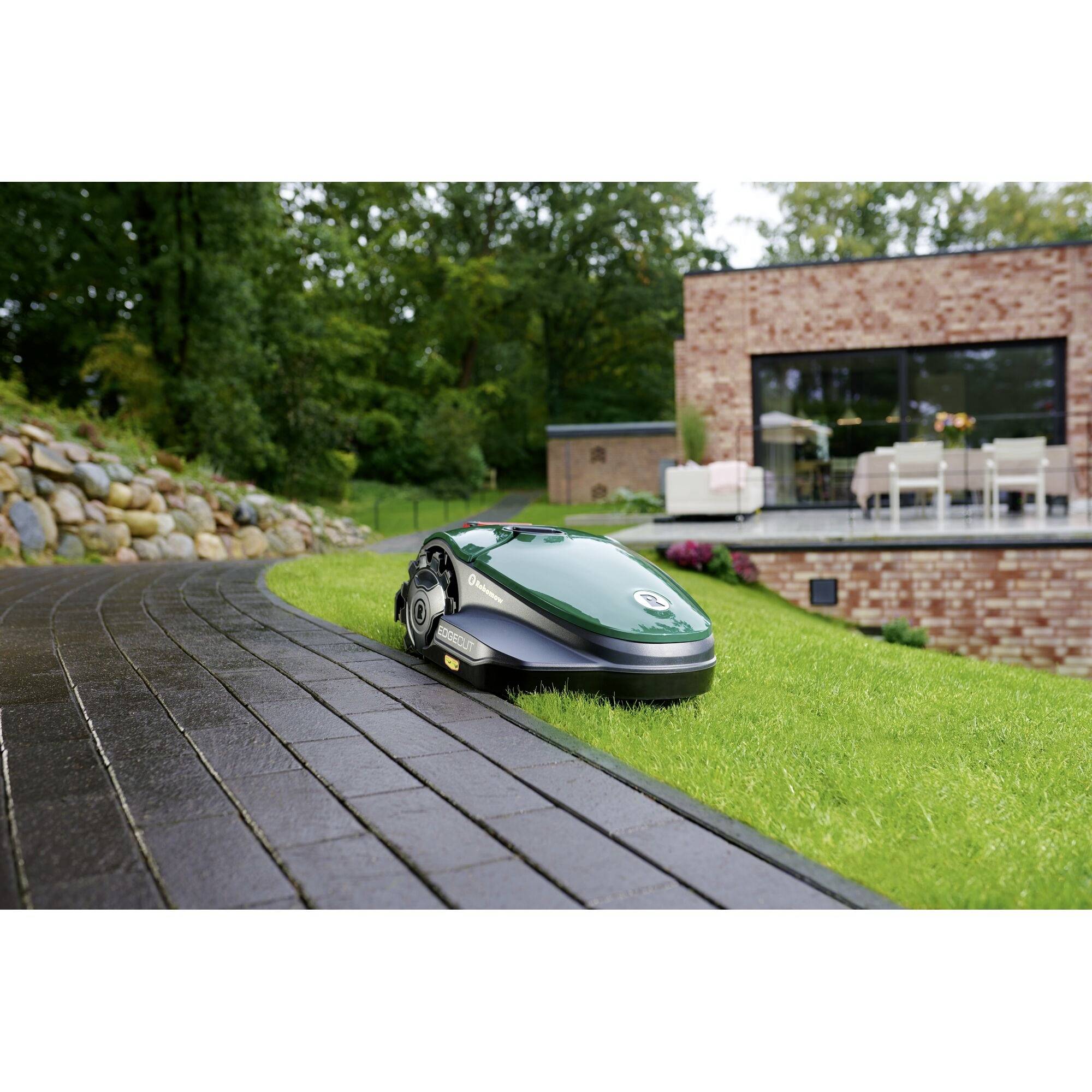 A green robotic lawnmower operates on a lawn beside a patio with modern furniture, surrounded by trees and a brick house.