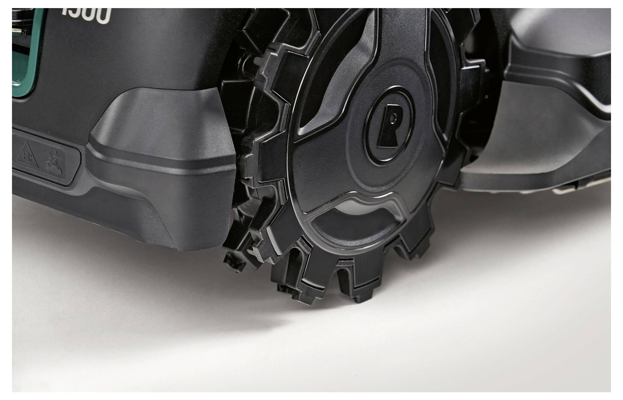 Close-up of a robotic lawnmower, focusing on one of its black, serrated wheels against a grey background.