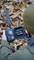 A person is holding an open trail camera device with a battery compartment and screen, surrounded by autumnal leaves in the woodland.
