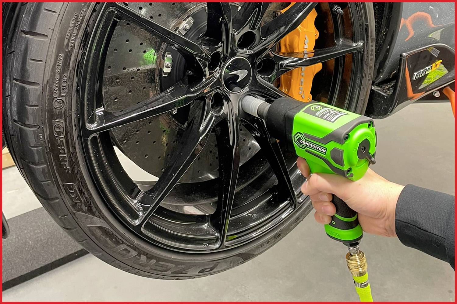 A person is tightening the wheel nuts of a car with a green impact wrench. The wheel has black rims and orange brake callipers.