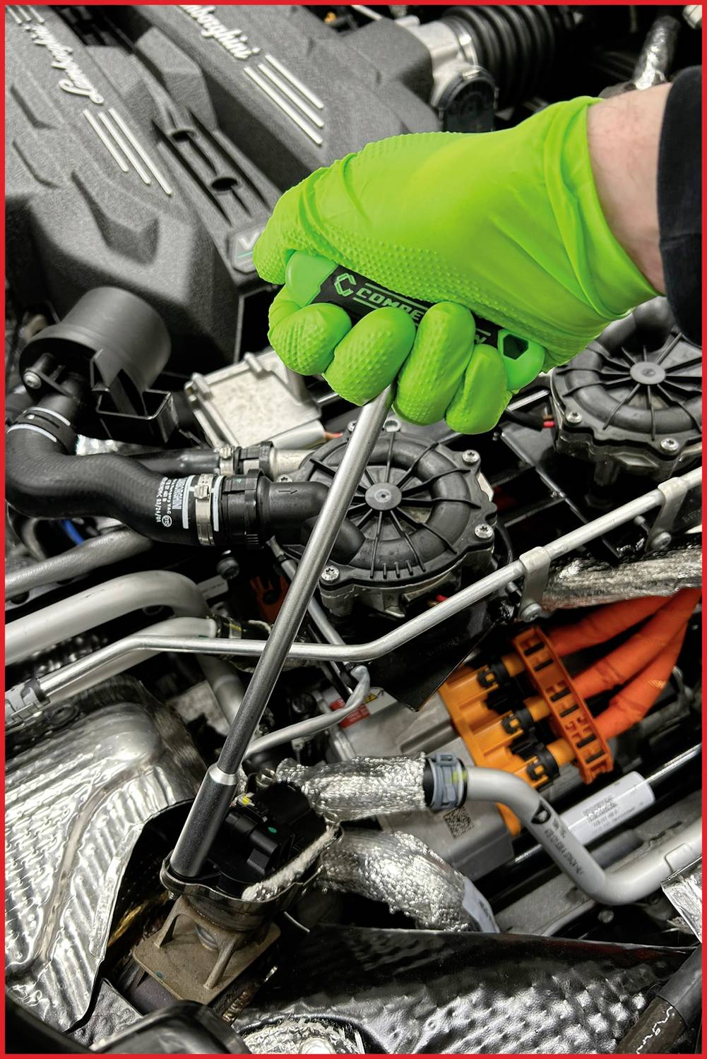 A person wearing green gloves is working with a spanner on an engine. Visible cables and hoses are present in the engine compartment.