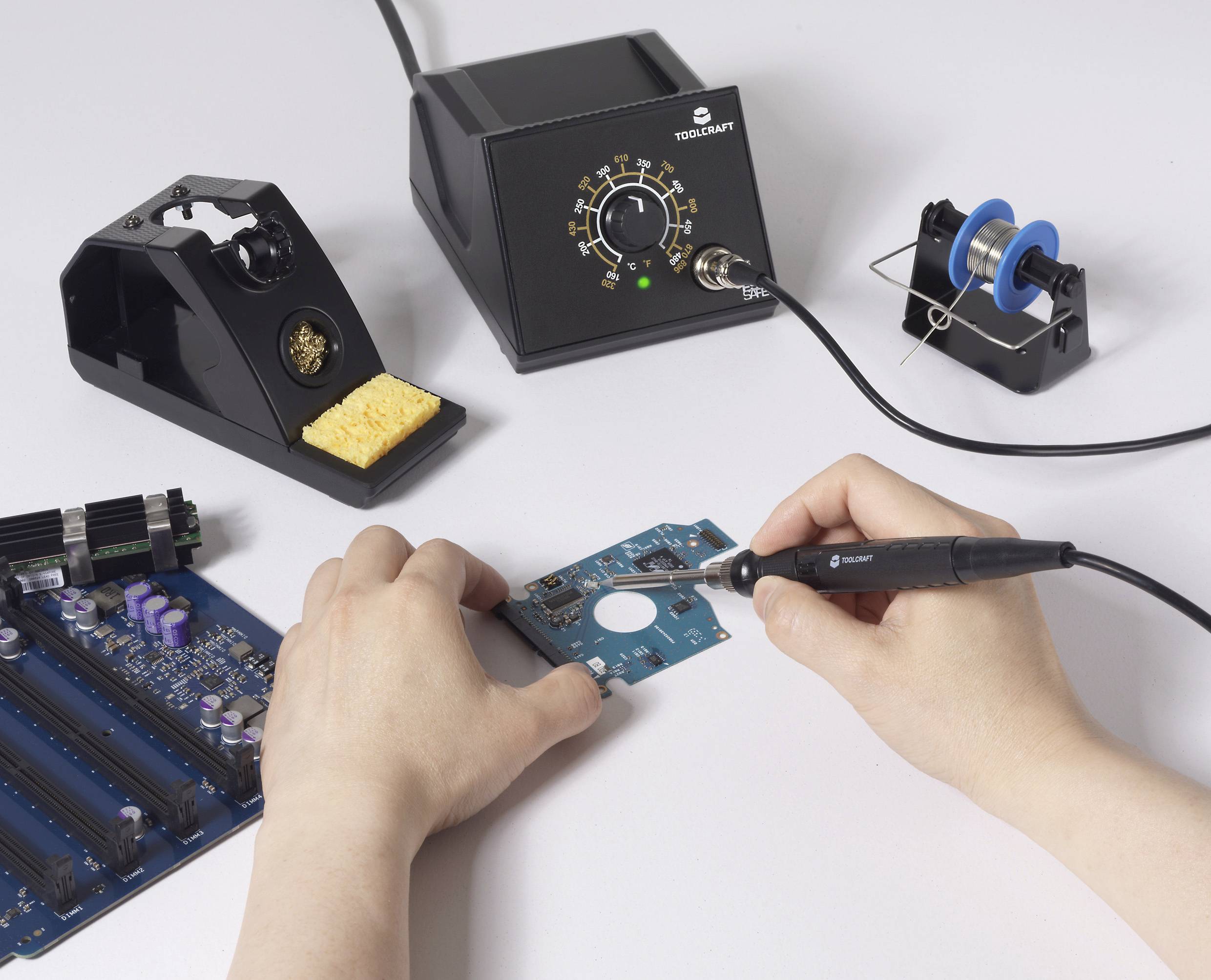 A person soldering electronic components on a circuit board using a soldering iron, with a soldering station and spool nearby.