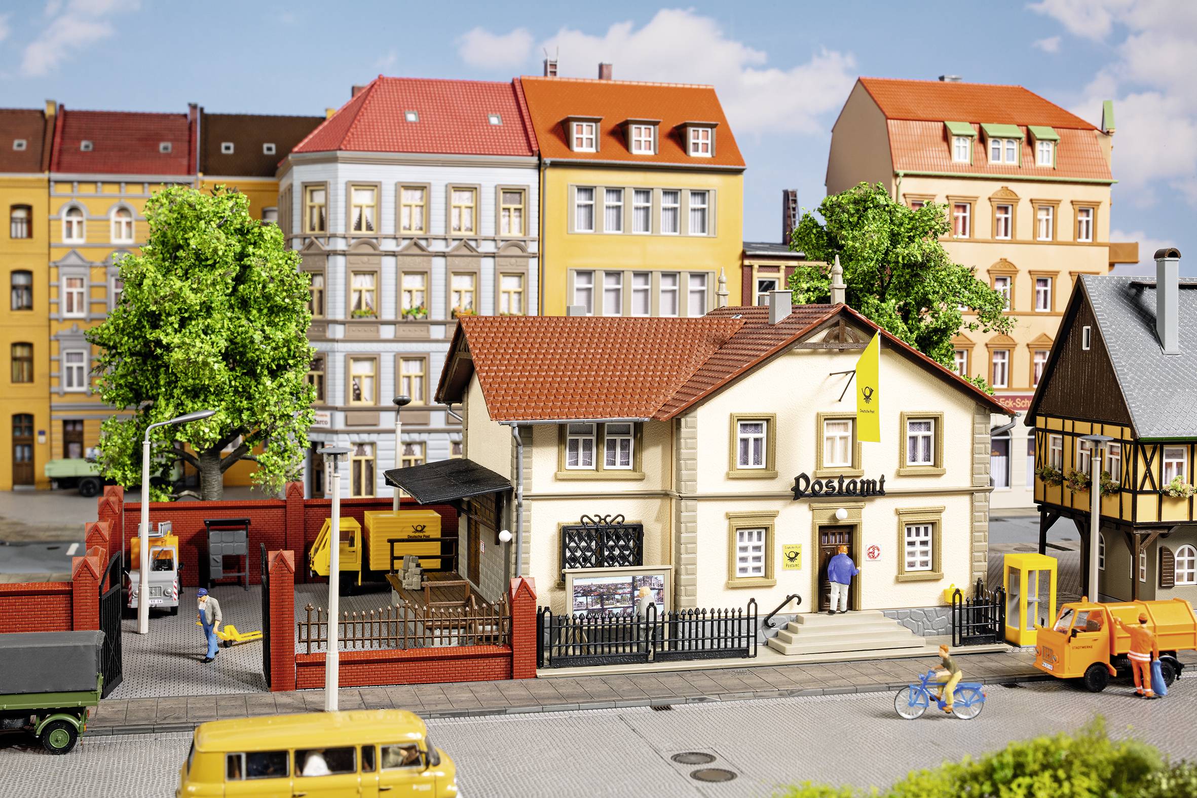 A miniature village with colourful buildings, a post office, trees and vehicles. One person is cycling on the street.