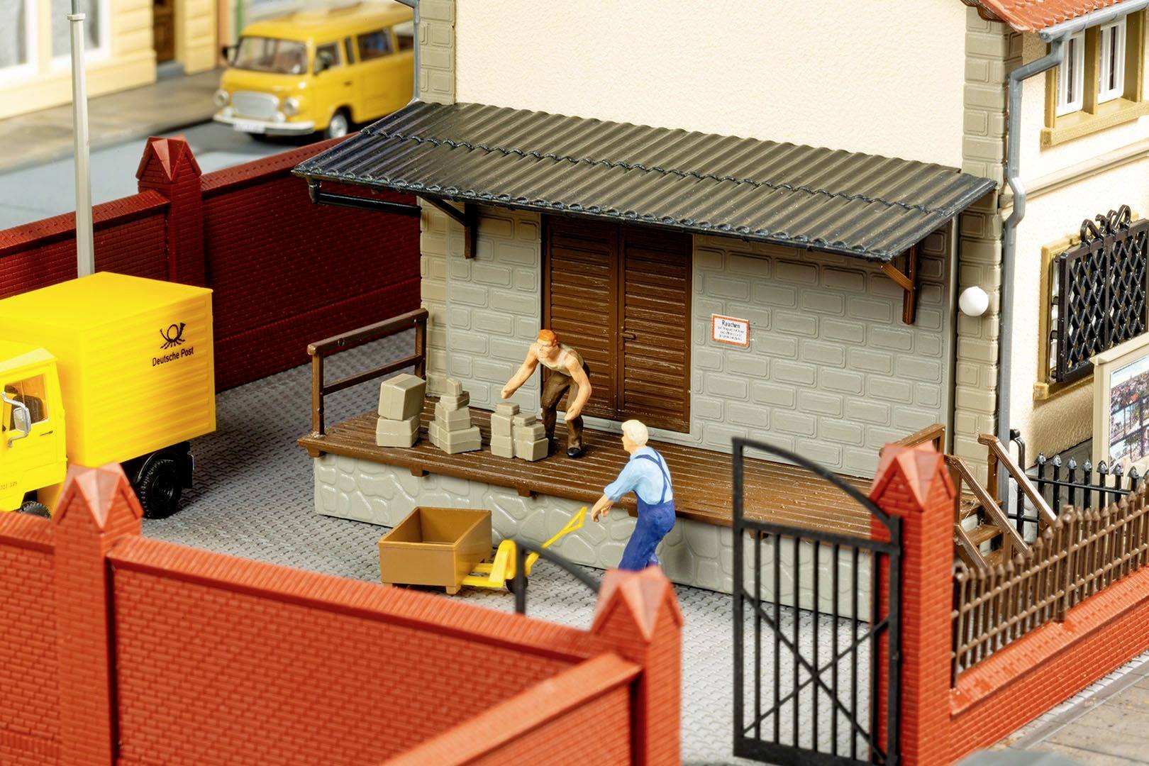 Two miniature figures are unloading boxes on a small yard in front of a building with a red fence. A yellow delivery van is parked on the left.