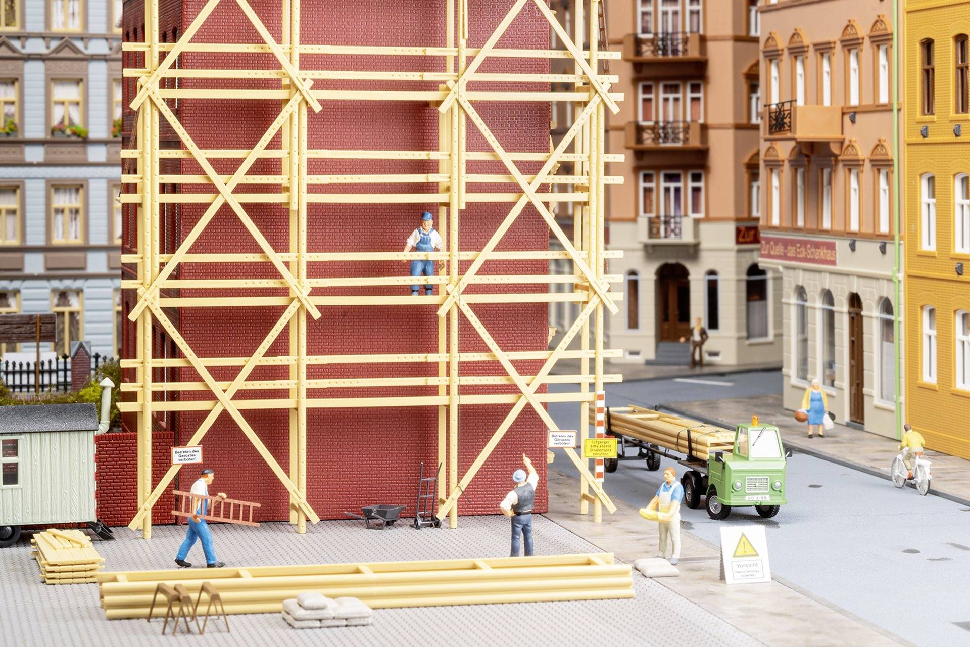 A miniature scene depicts workers on scaffolding in front of a building on a city street. Wooden planks and construction tools are scattered around them.