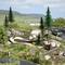 A detailed miniature model depicts a mountain bike park with a cyclist on a winding path, surrounded by trees and signs, near a car park with vehicles and hills in the background.