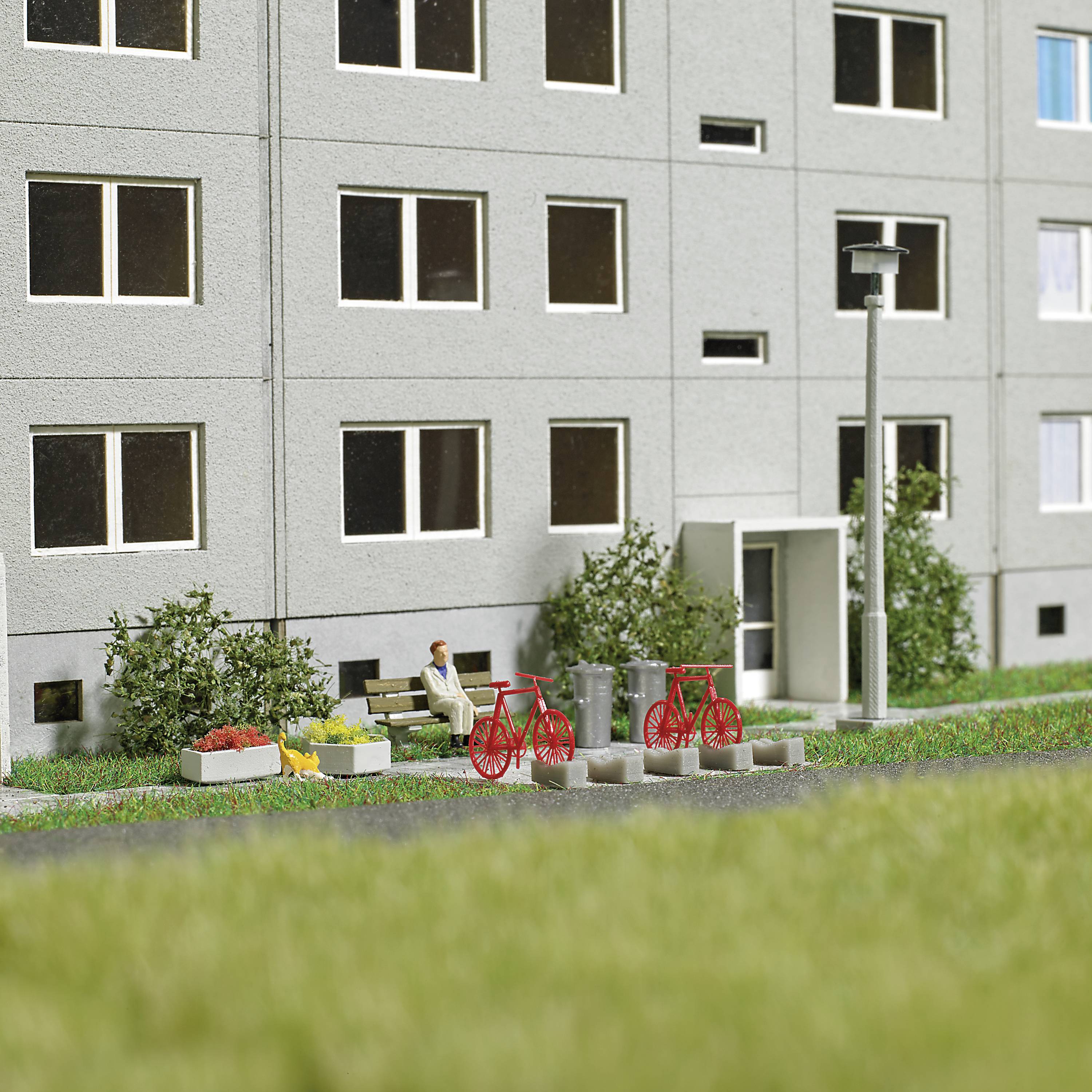 Multi-storey residential building with a grey facade, with red bicycles and benches in front, surrounded by bushes and a green lawn.