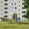 Housing block with multiple windows, green spaces and playground in the foreground. A few people can be seen on the playground.