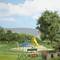 A playground with a slide and climbing frame next to a tree. In the foreground is a bench; in the background are hills and a meadow.