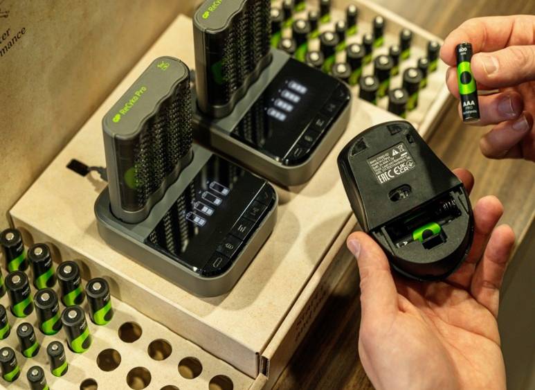 A hand is holding a battery compartment and inserting a rechargeable battery, with a charger featuring multiple charged batteries visible in the background.