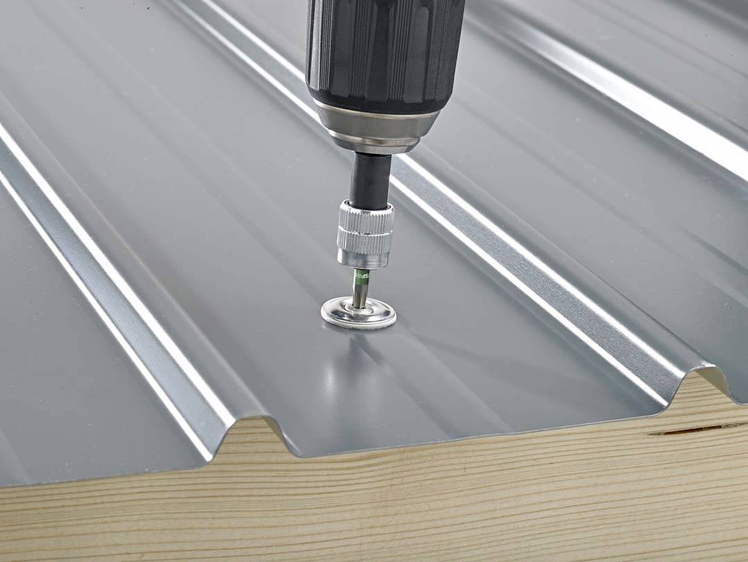 A cordless drill is screwing a self-tapping screw into a corrugated metal roof on a wooden substructure.