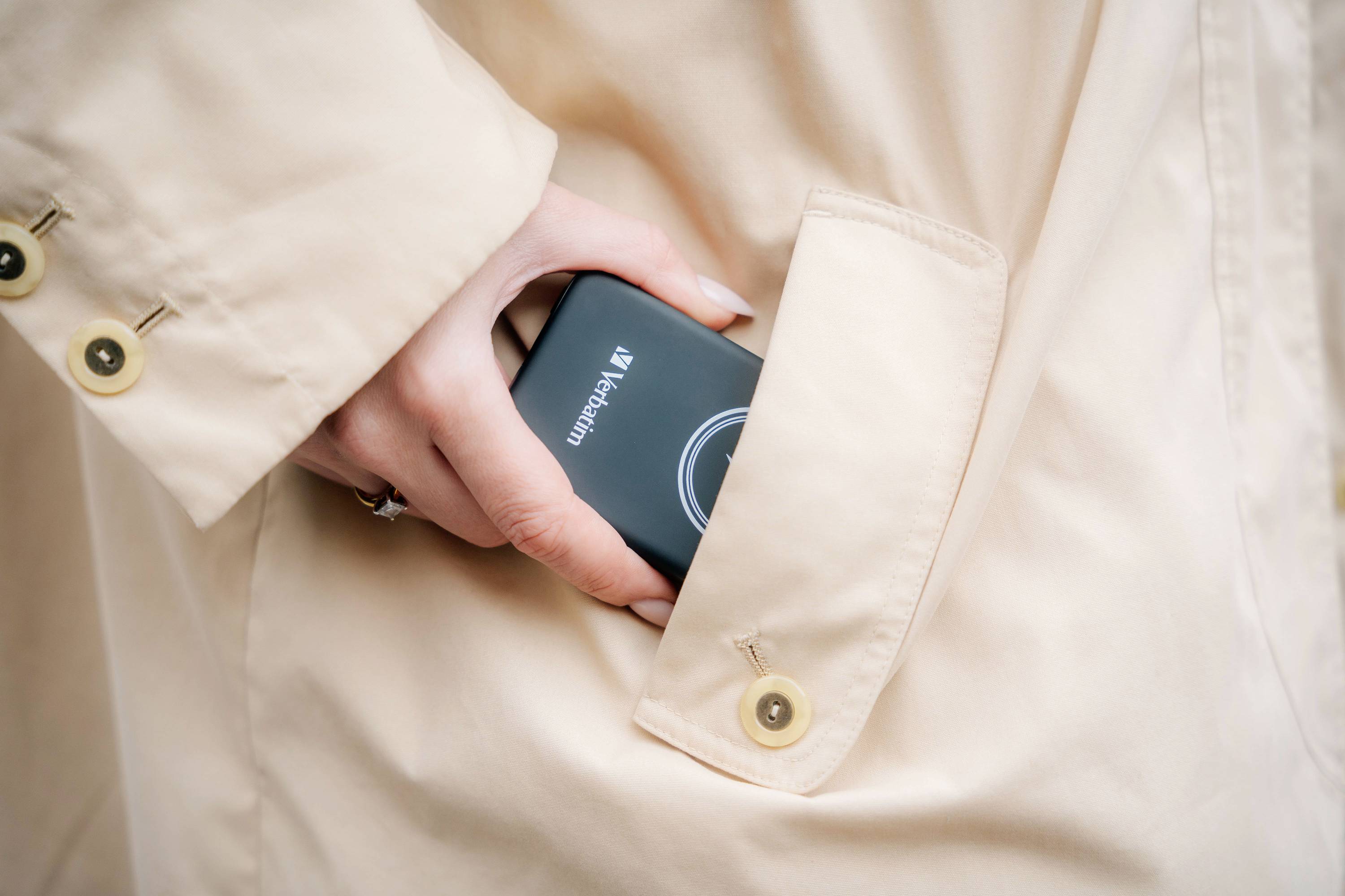 A hand is placing a portable hard drive into a beige coat pocket. The hard drive is black and displays the brand logo.
