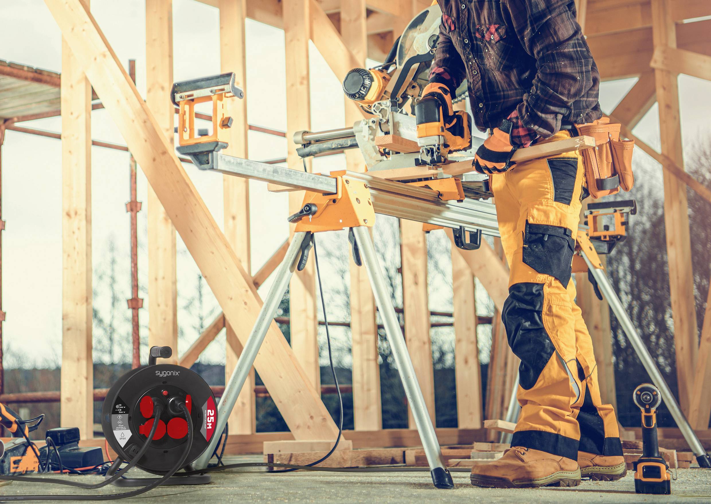 'A person in safety gear working with a miter saw on a construction site, framed by unfinished wooden structures. Equipment nearby.'