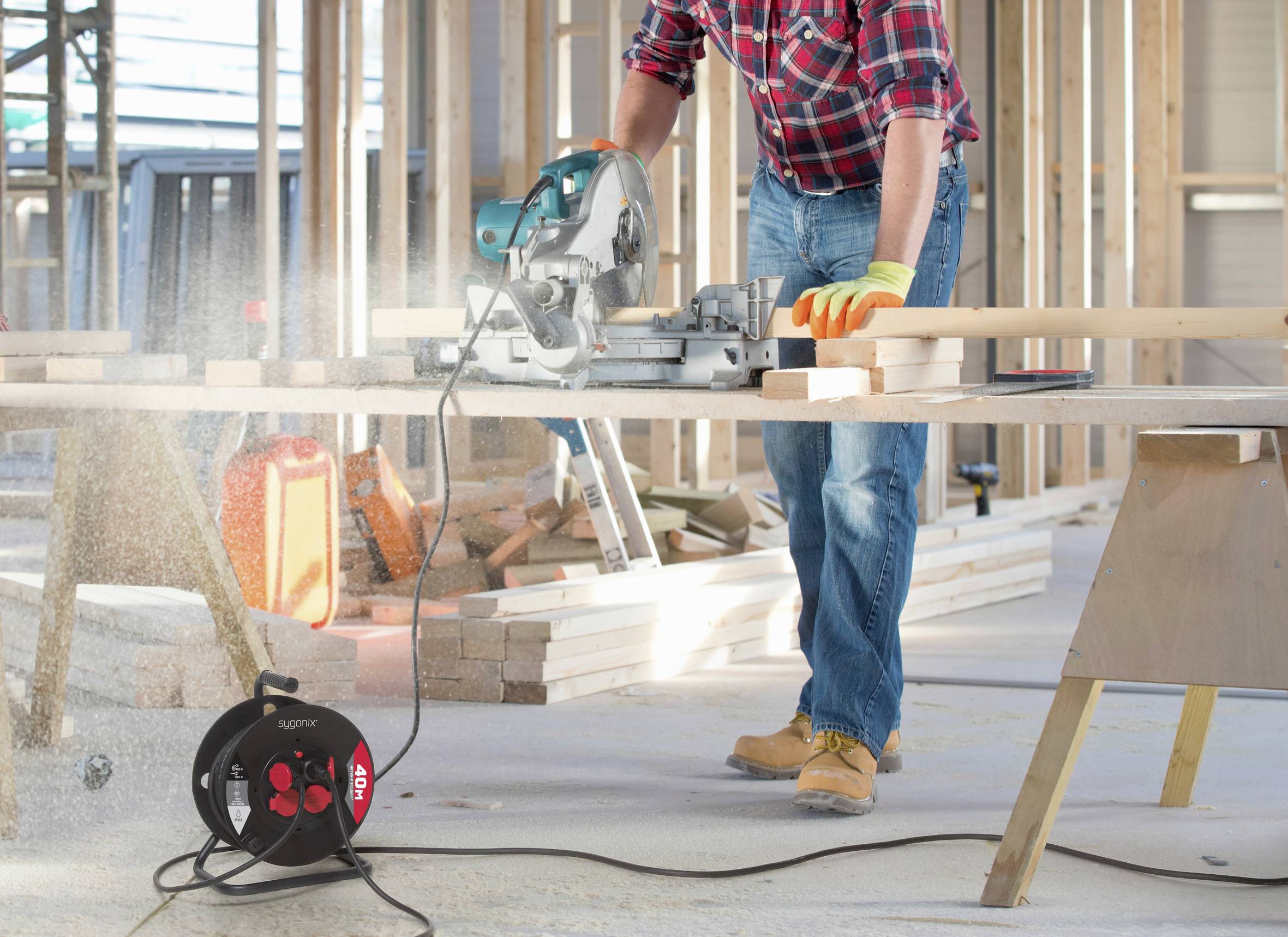 A person in work attire uses a circular saw on wooden beams in a construction site, surrounded by wood and construction tools.