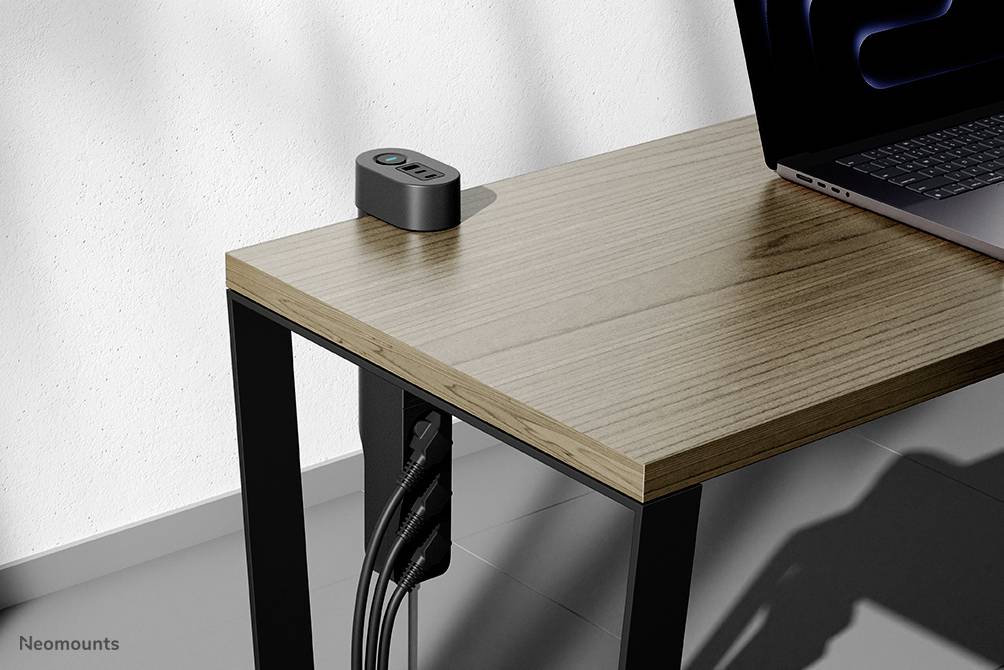 A modern wooden table with black metal legs. A small grey device with a blue button sits on the table. A laptop is partially visible. Cables run neatly along one of the table legs.