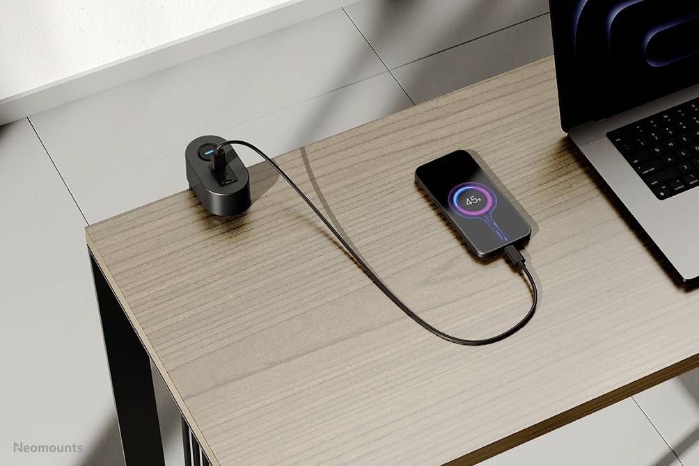 A smartphone on a table next to a laptop, connected via a charging cable to a power socket. The display shows 65% battery level.