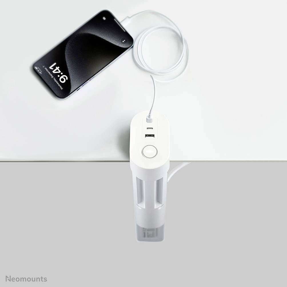 A smartphone is connected to a desk charger that is attached to the edge of the table. The charger has two USB ports.