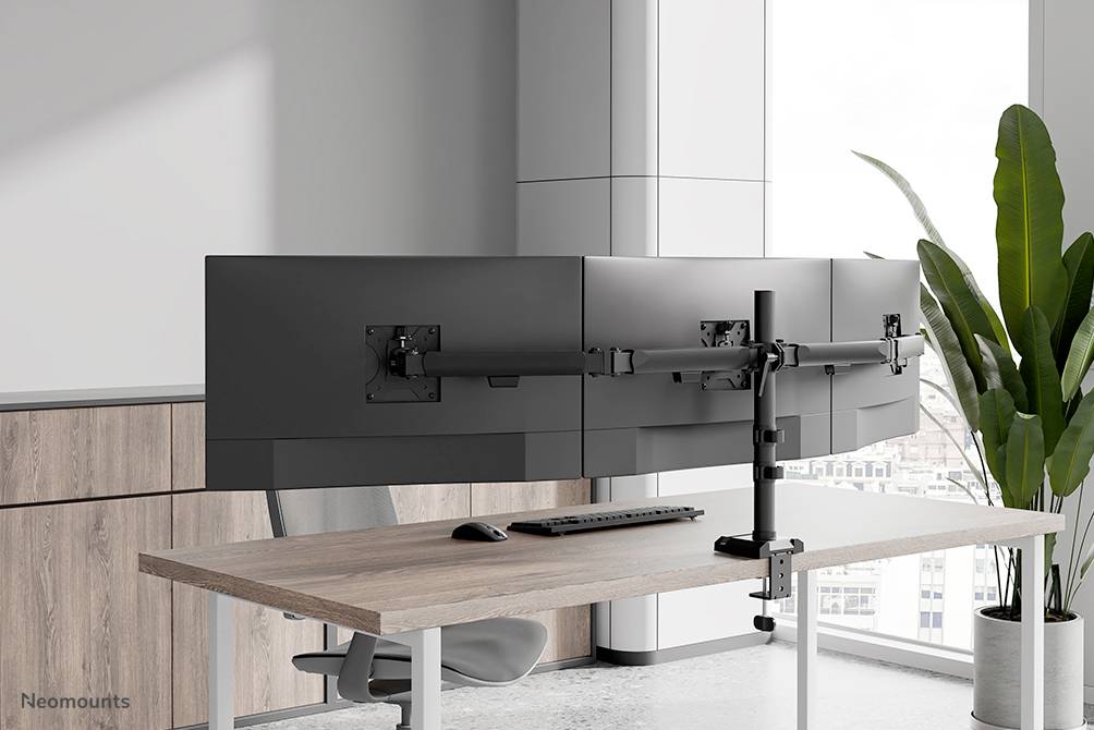 A modern office workstation with four monitors mounted on a desk arm. The room is bright and minimalist with large windows.