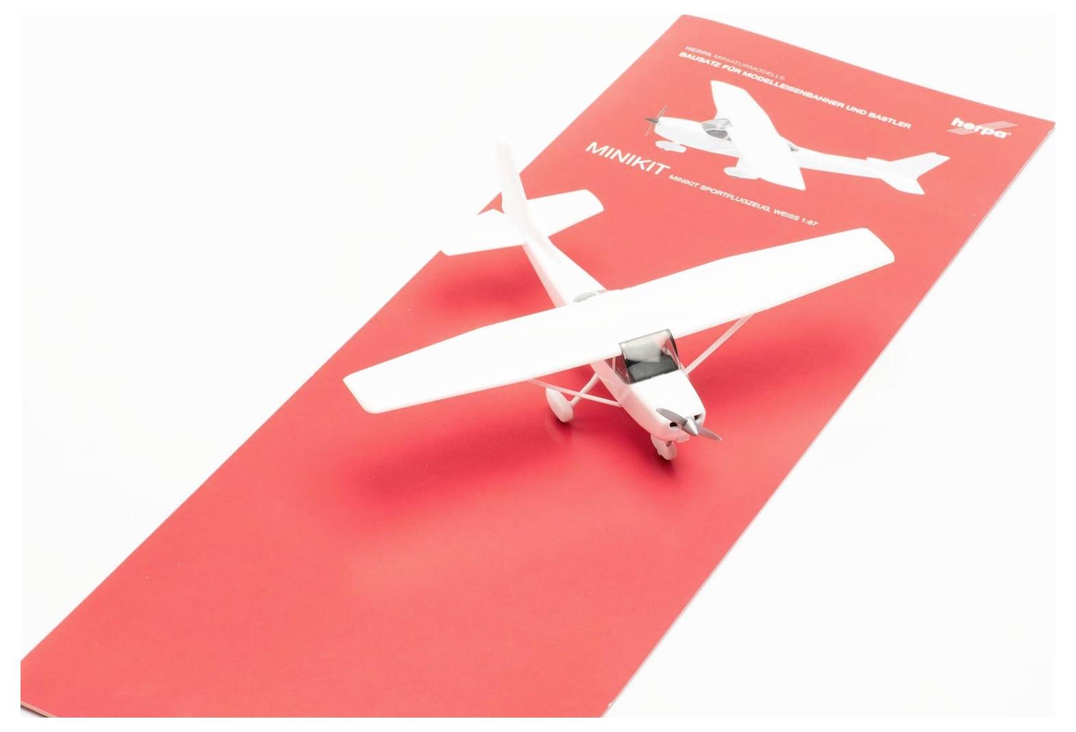 A white model airplane on a red package with the inscription 'Minikit'. The airplane is positioned diagonally, with the front facing left.