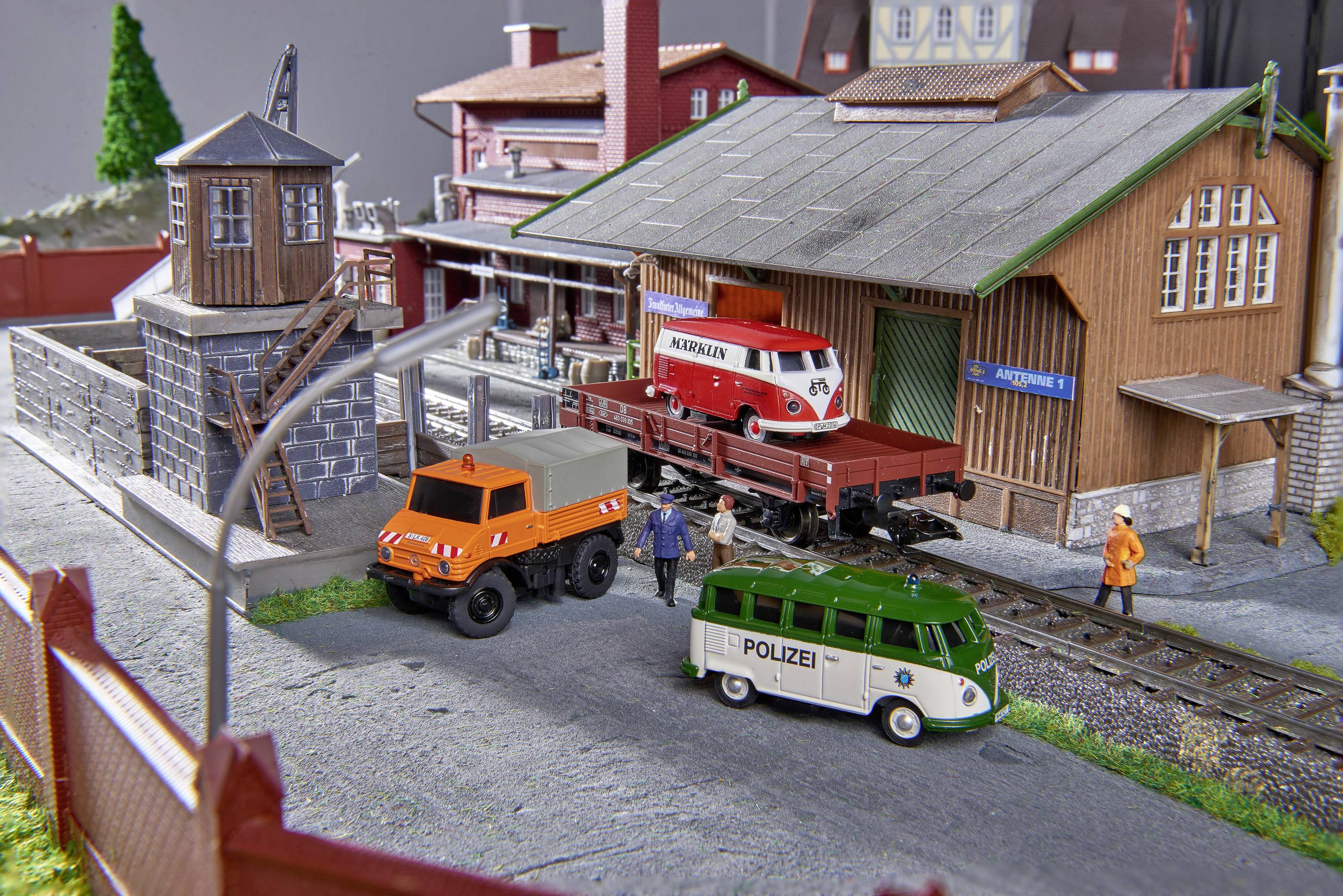 Model railway scene with multiple vehicles: An orange lorry, an ambulance, and a green police van near a goods shed. Two figures stand as workers on the site.