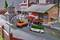 Model railway scene with multiple vehicles: An orange lorry, an ambulance, and a green police van near a goods shed. Two figures stand as workers on the site.