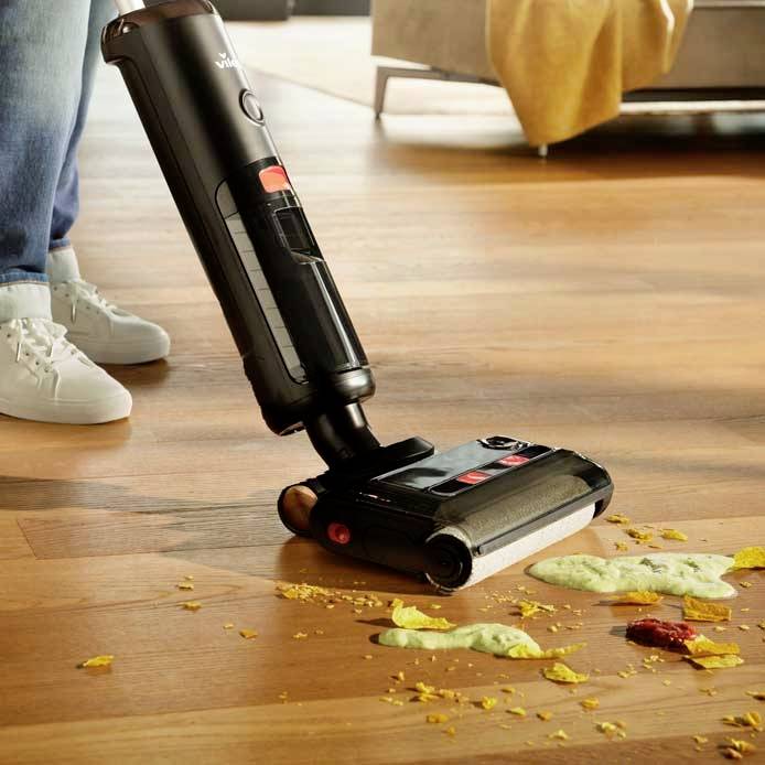A cordless vacuum cleaner simultaneously sucks up crisps and pudding from a wooden floor, while someone in white trainers stands nearby.