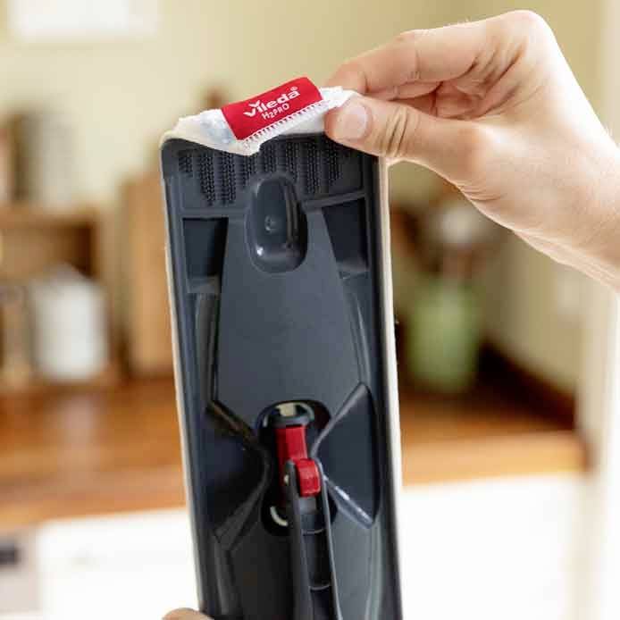 A hand attaches a white cleaning cloth to a mop with a red Vileda logo. A blurred kitchen is visible in the background.