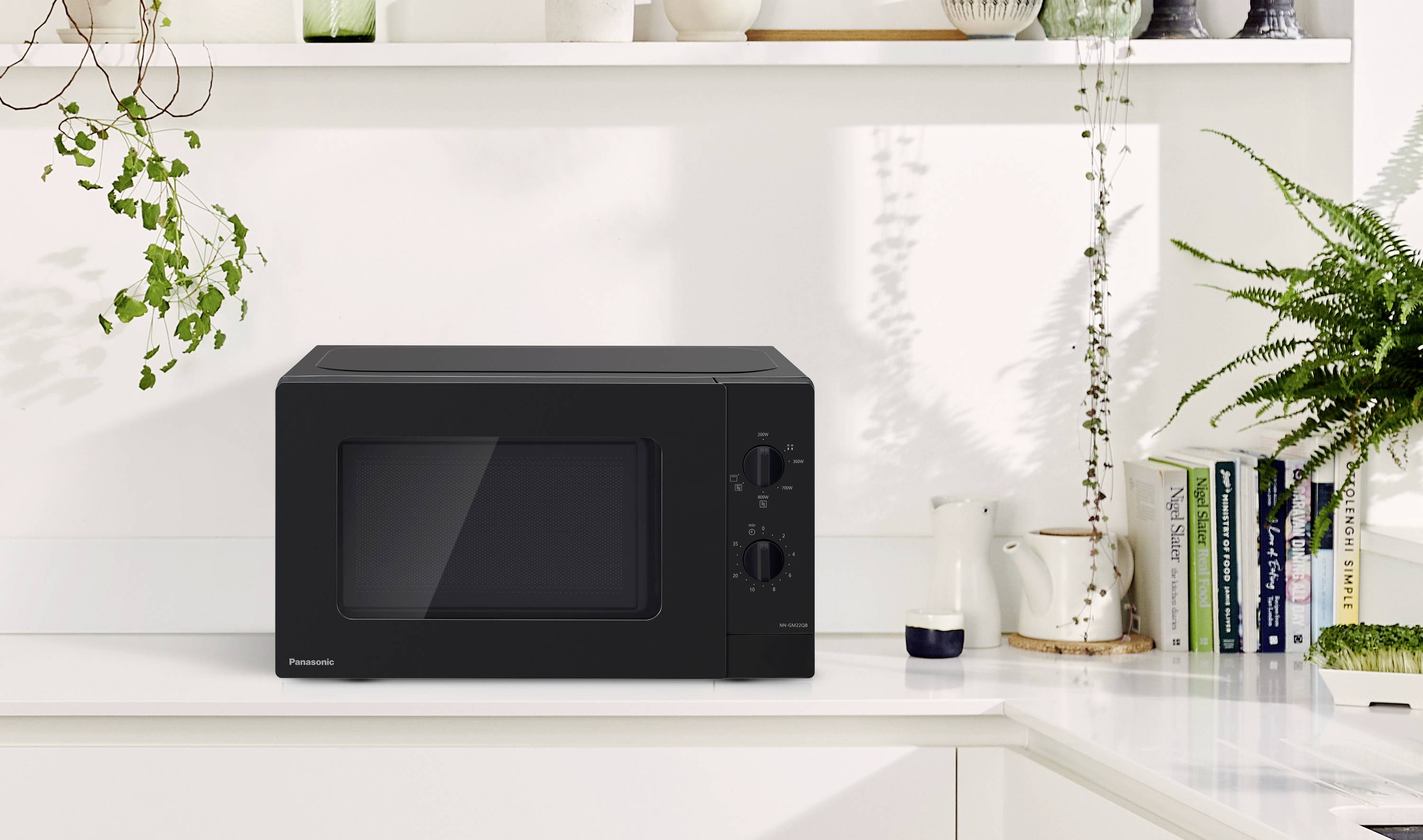 Black microwave on white kitchen worktop, surrounded by plants and crockery. Shelves with books in the background.