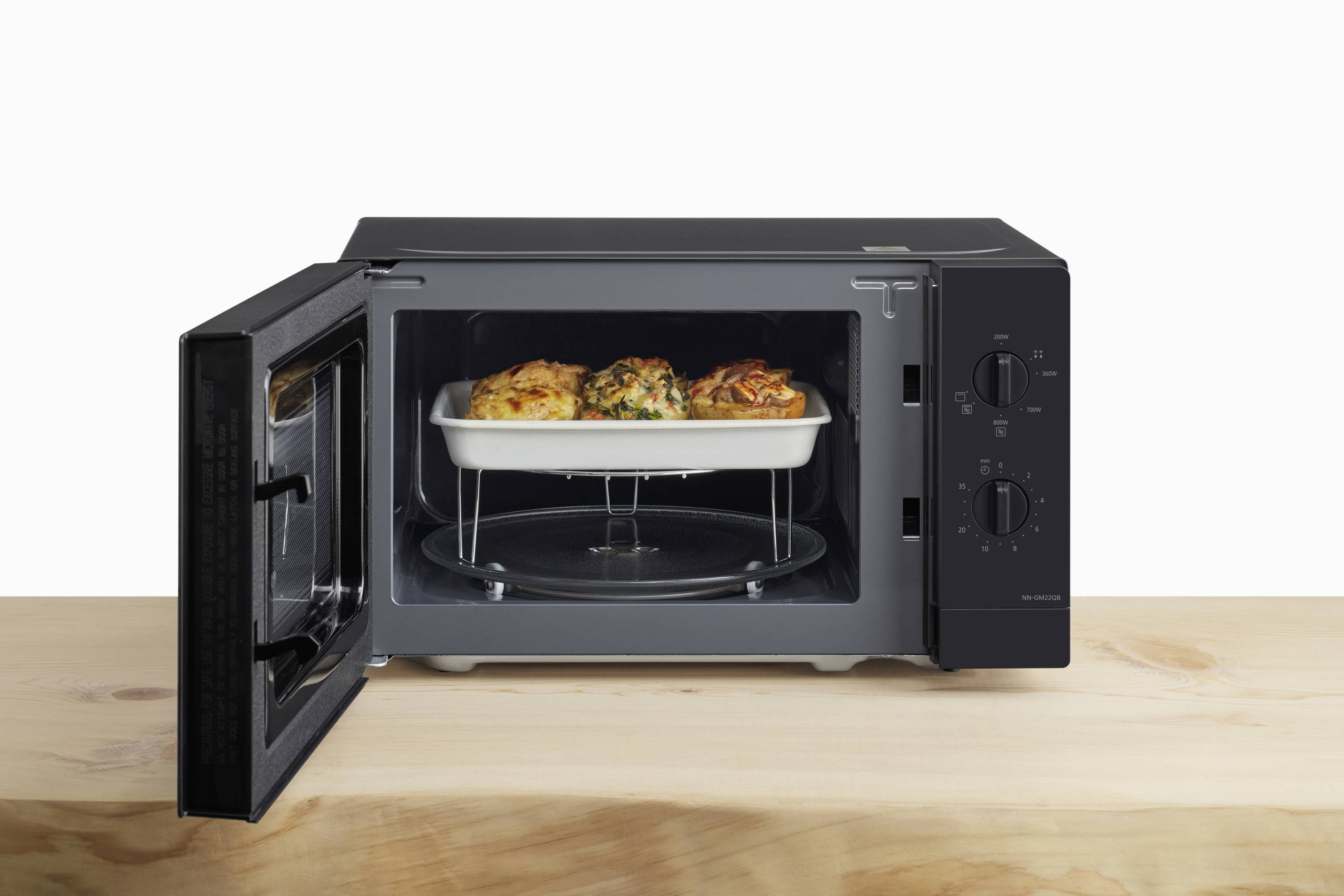 A black microwave oven on a wooden table with its door open reveals a baking dish containing three gratinated dishes inside.