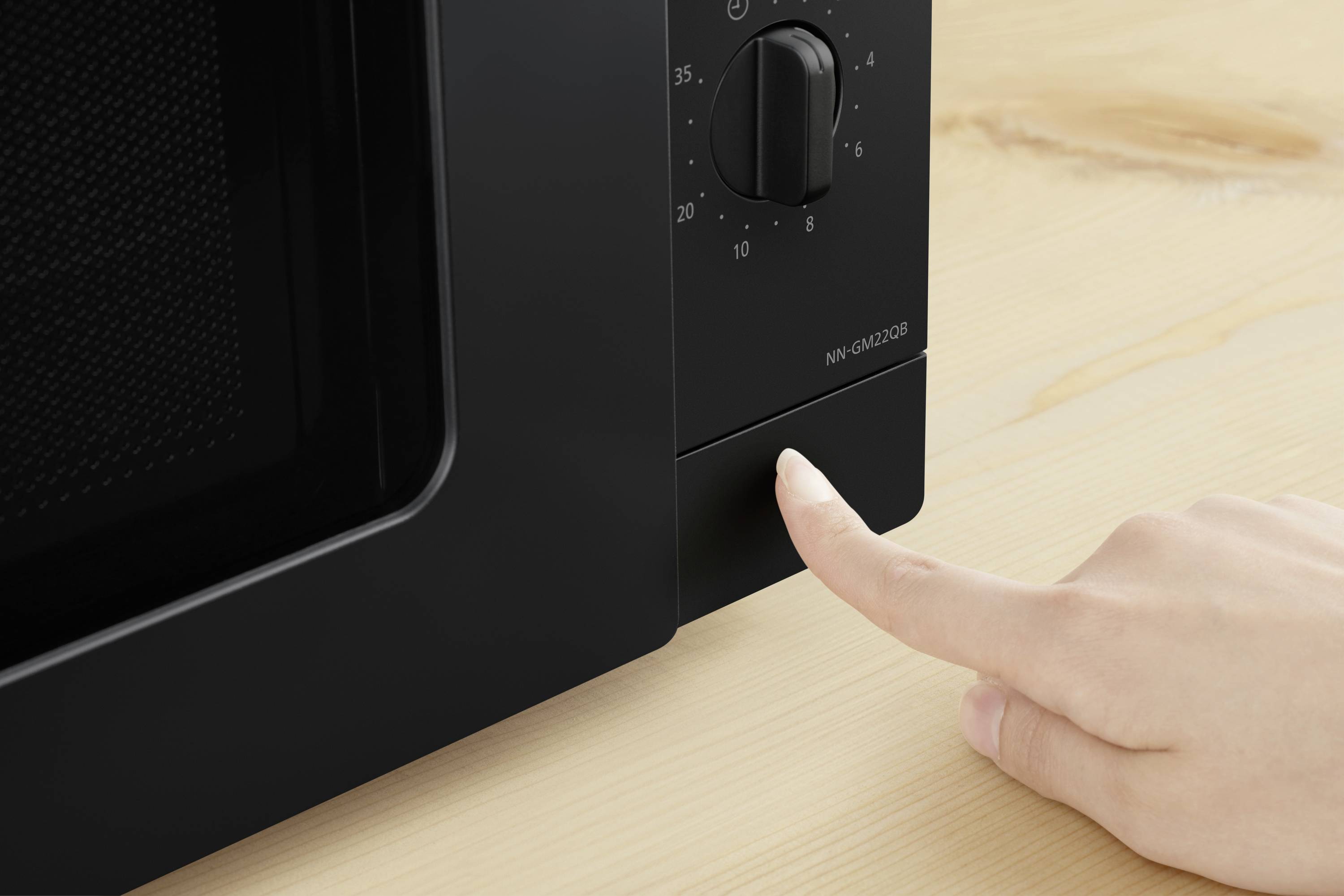 A hand presses the button of a black microwave oven on a wooden surface.