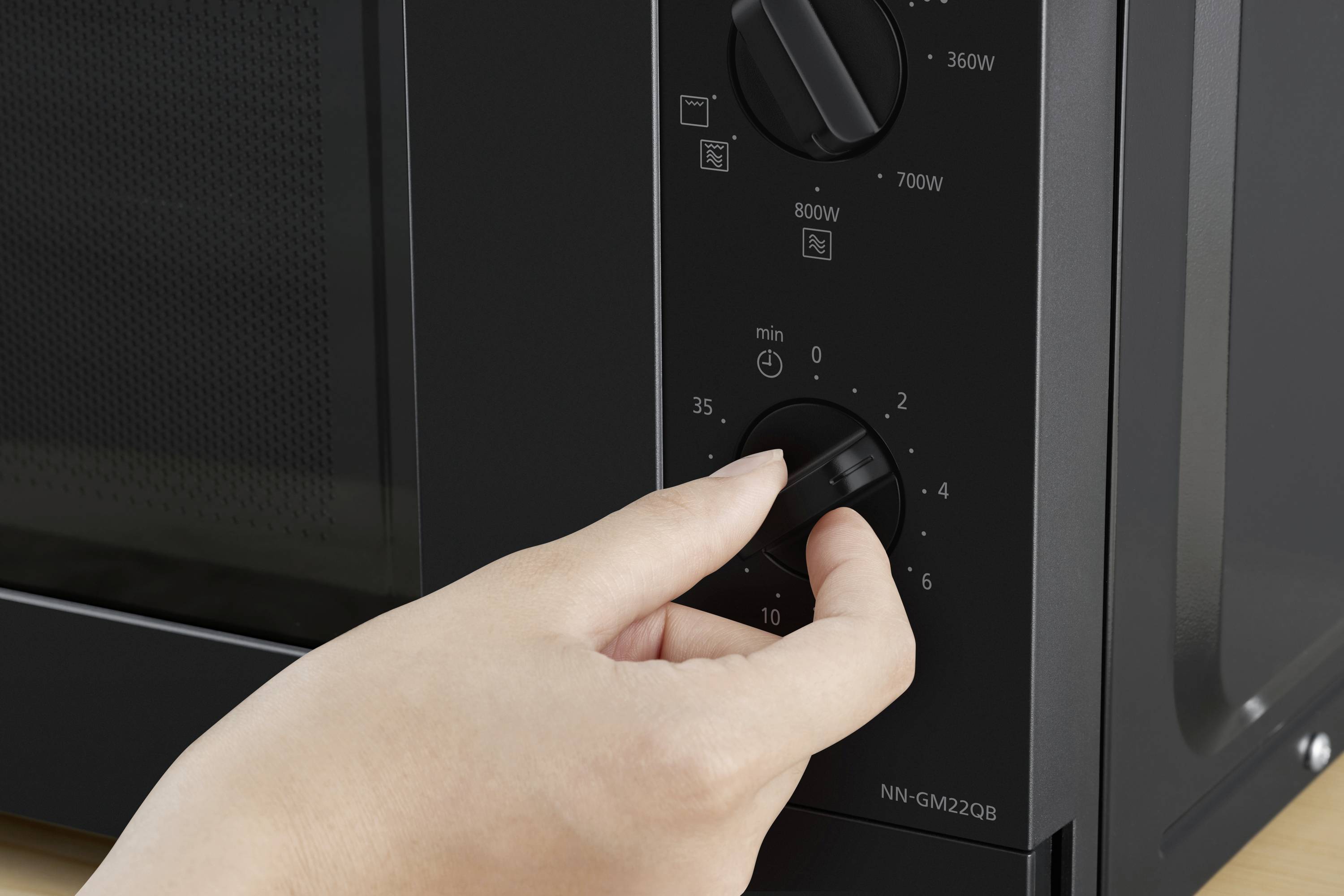 A hand turns the timer knob of a microwave. The scale shows 'min' and numbers up to 35, with power settings of 360W, 700W, 800W.