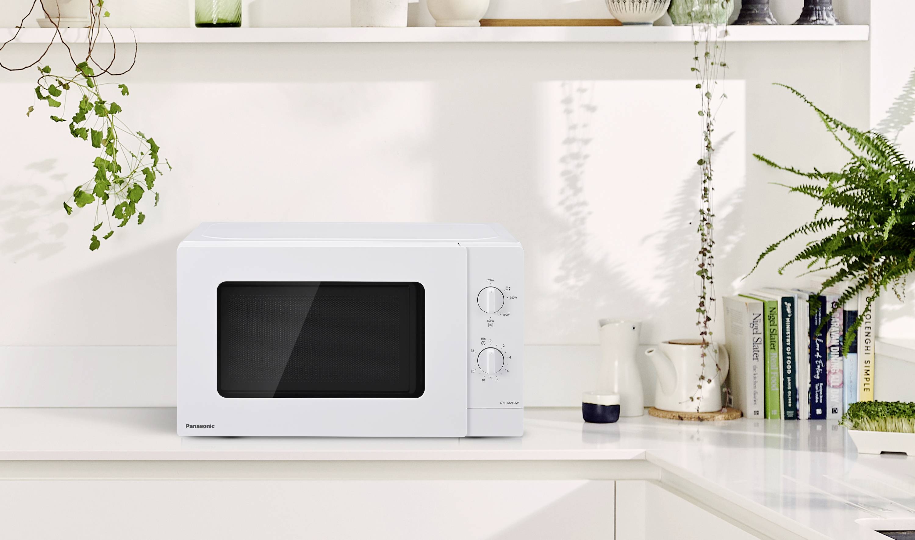 White microwave on a kitchen worktop, surrounded by plants and books.