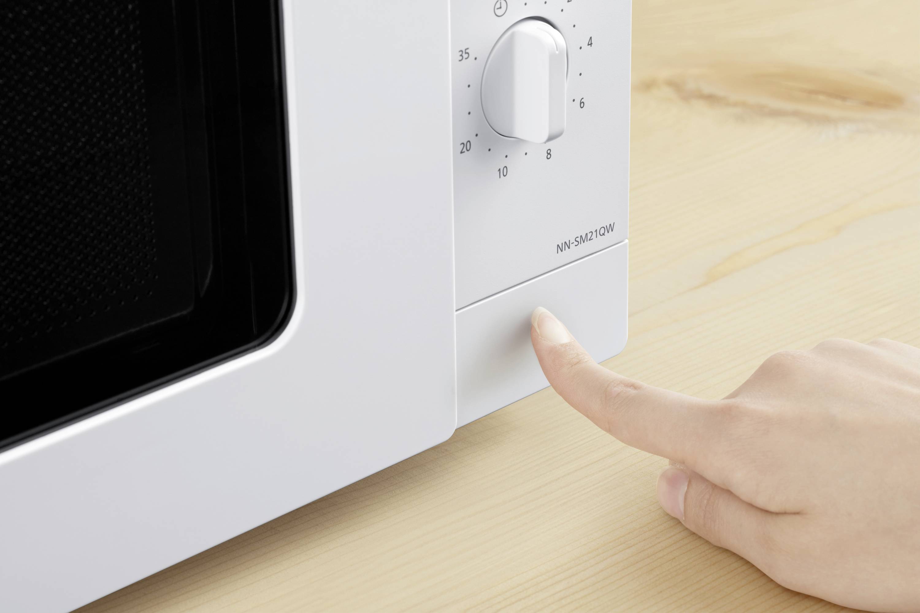 A hand presses a button on a microwave on a wooden table. The microwave has a rotary dial and a time scale.