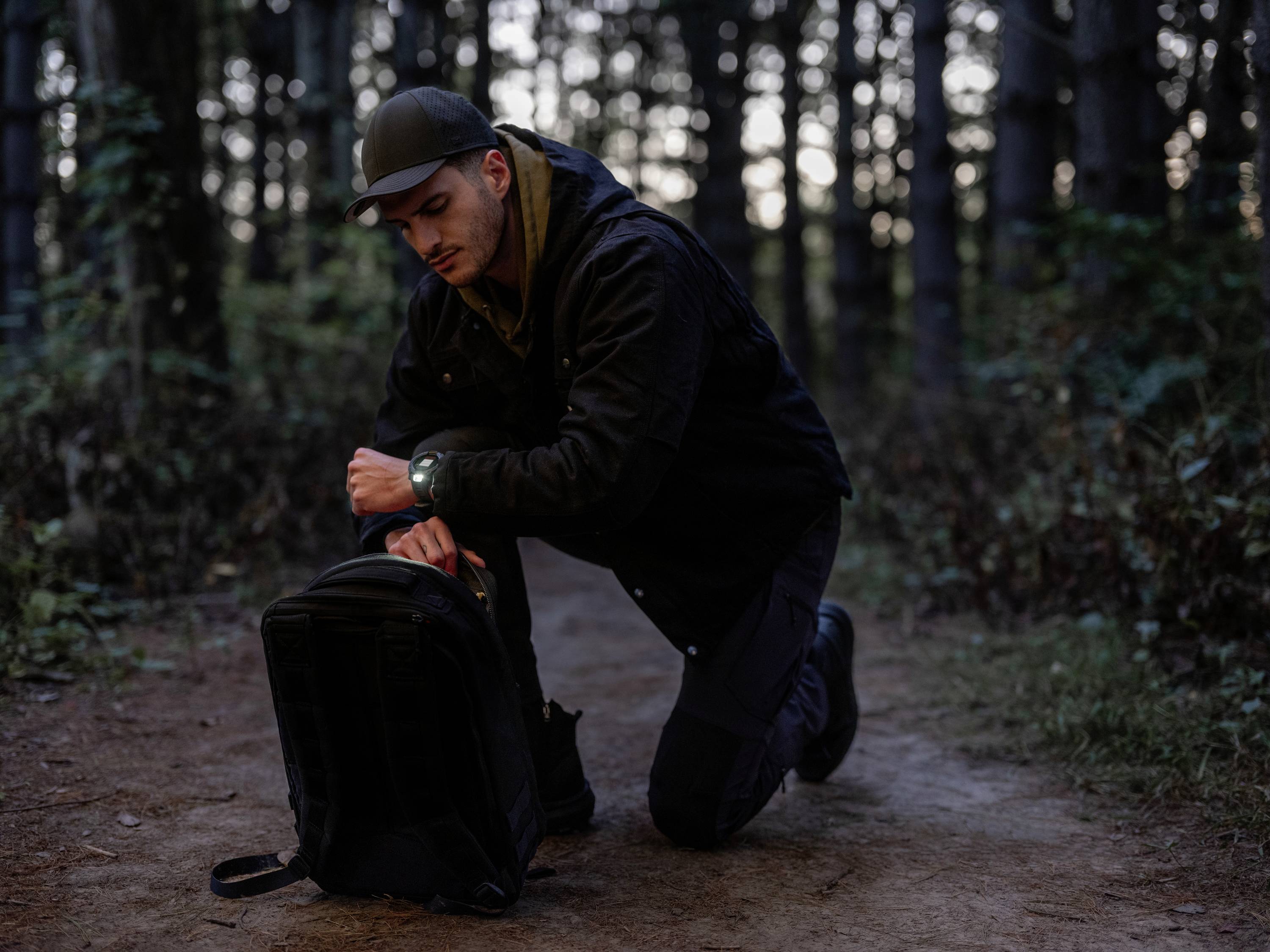 A person is kneeling on a forest path and opening a black rucksack. The surroundings are surrounded by trees, with a dim, shadowy atmosphere.