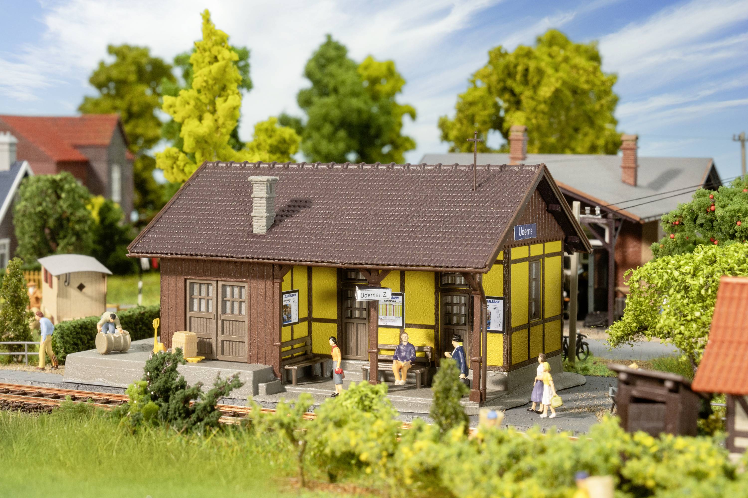 Small model of a railway station with a yellow building and tiled roof. People are sitting and standing in front of the building. Background: Trees.