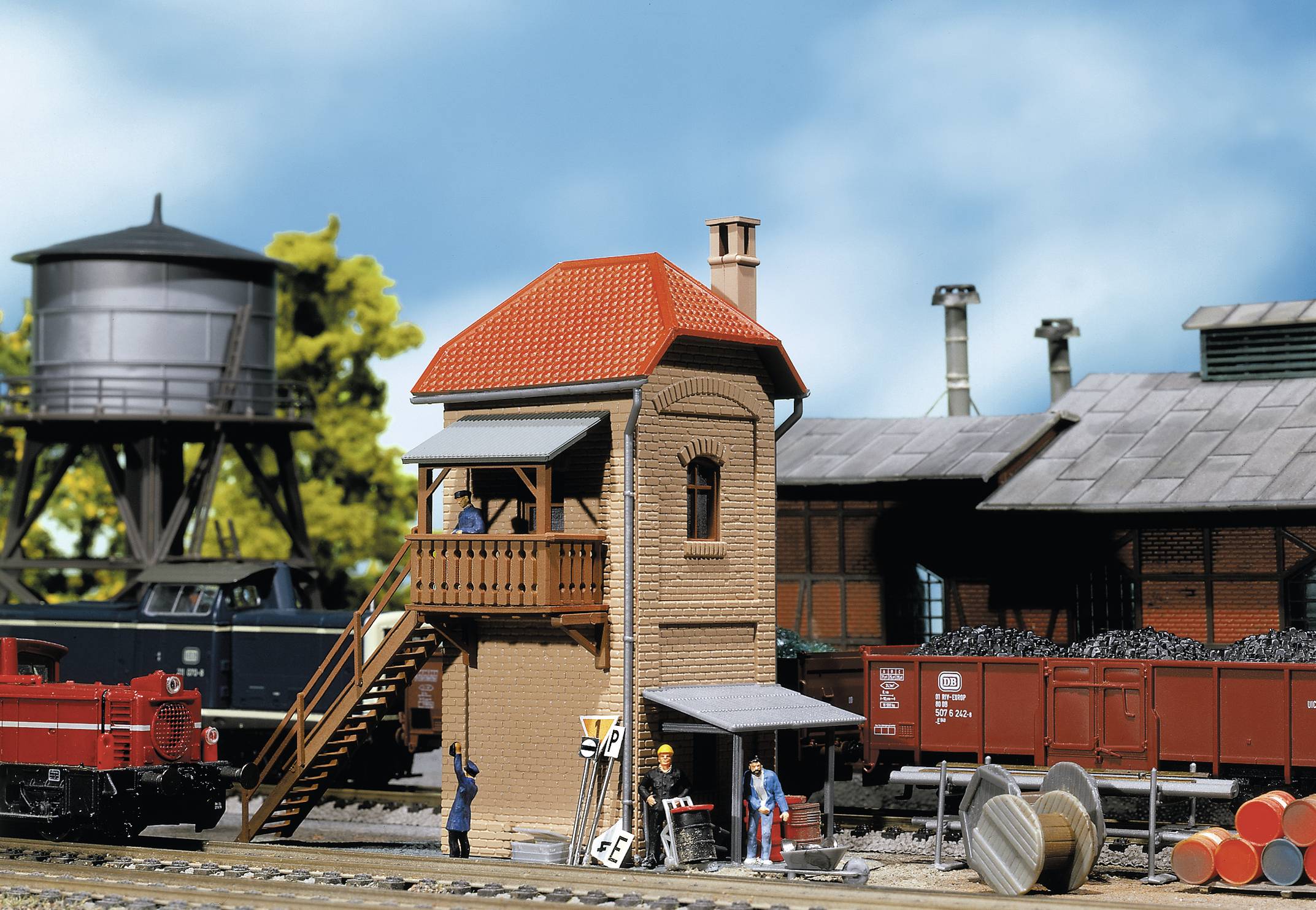 A model railway scene with a signal box, two locomotives and several people. In the background, a water tower and warehouse.
