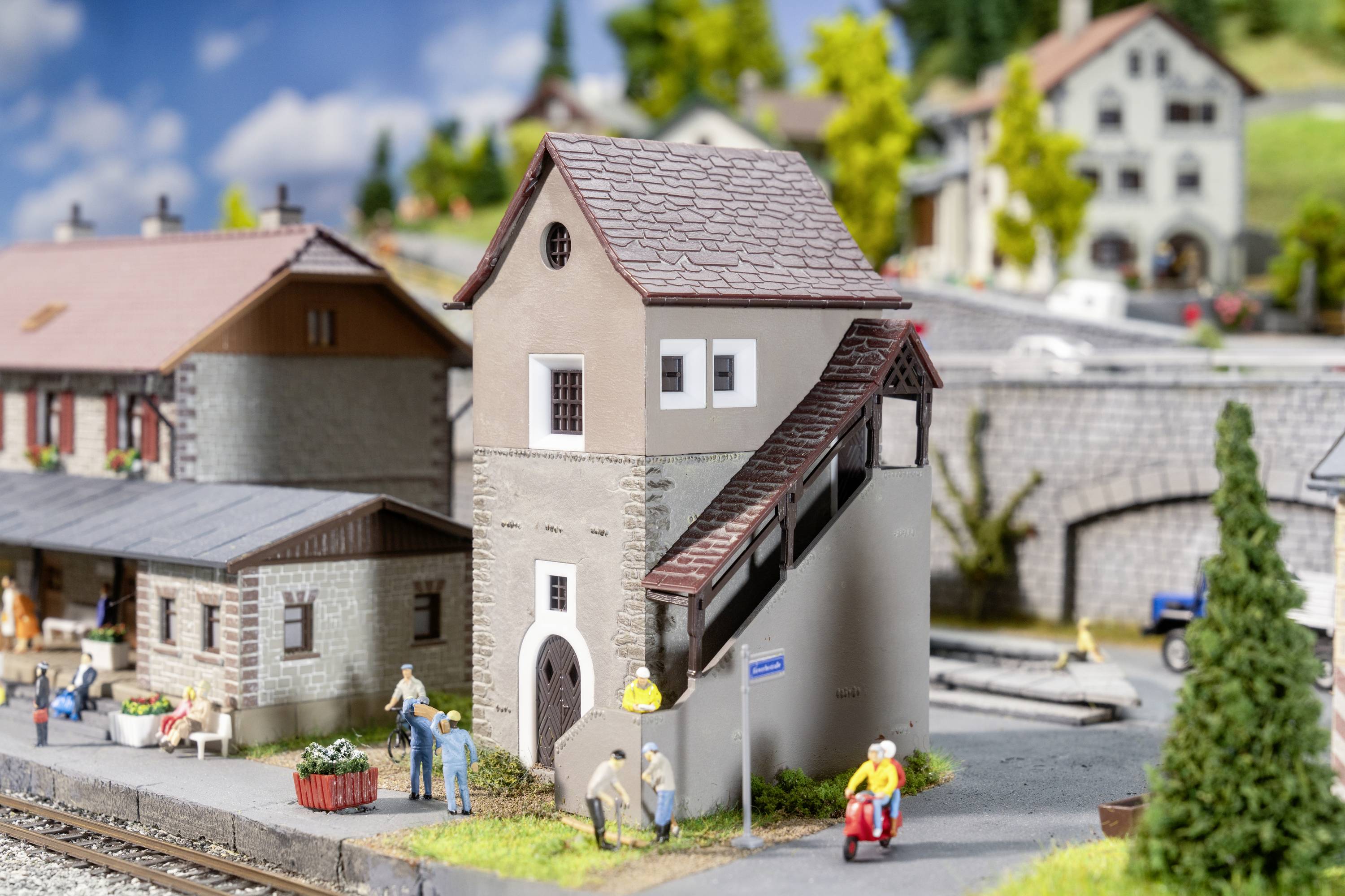 A model railway diorama depicts a village with miniature buildings, people, and trees. A tower with a staircase stands at the centre.