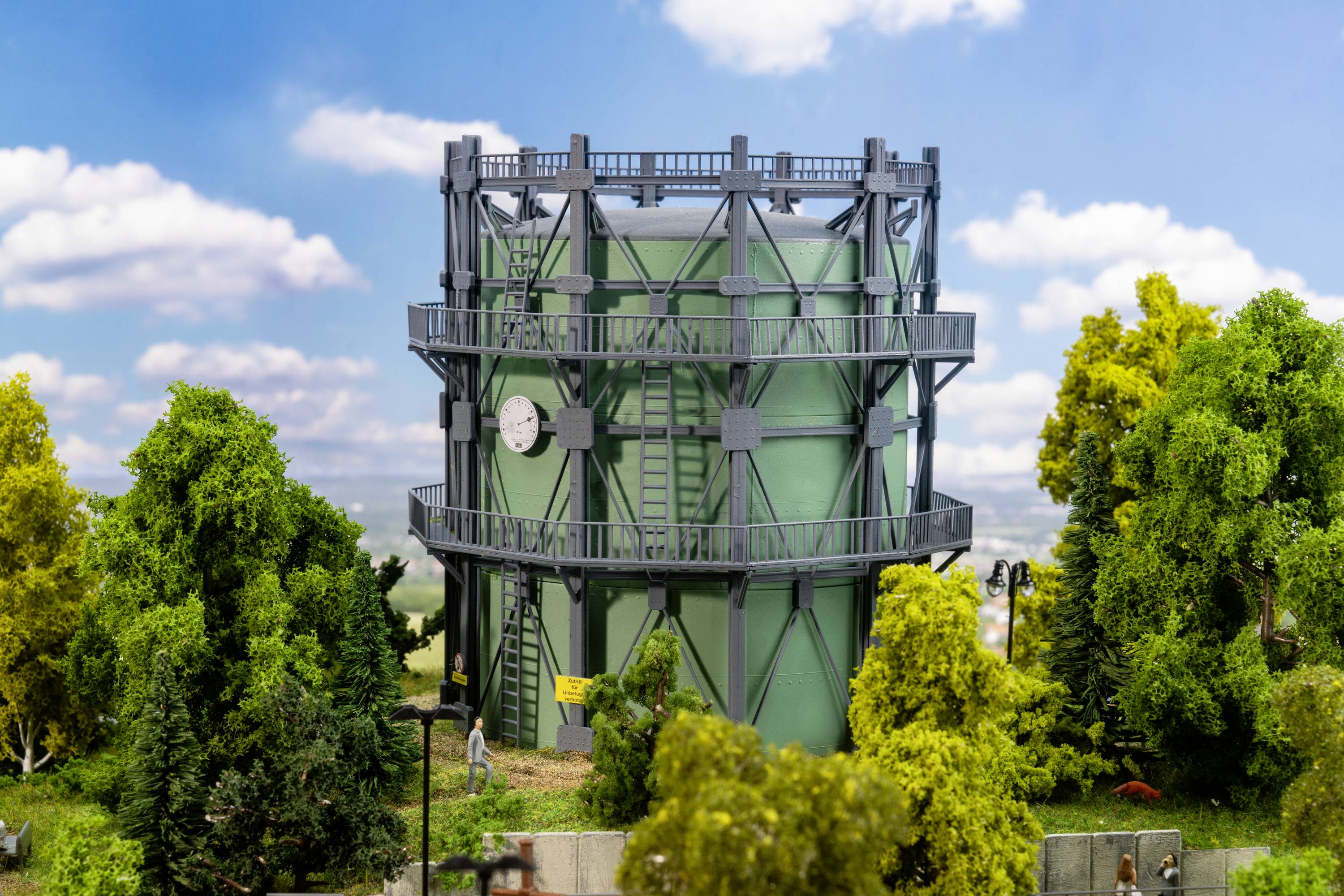 The image depicts a green gas storage tank with a steel frame in a rural setting, surrounded by trees and a blue sky.