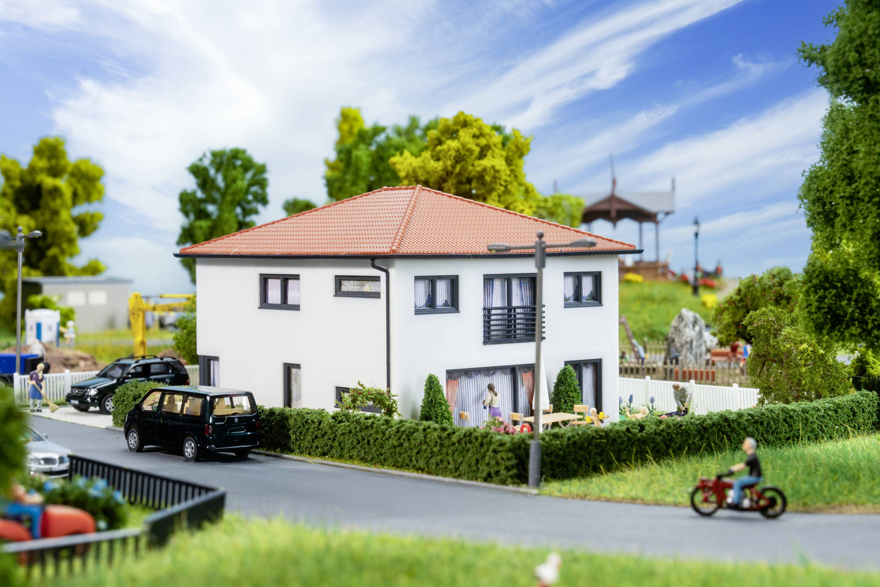 A modern two-storey white house with a red roof stands on a street, surrounded by trees and a garden. A pavilion is visible in the background.