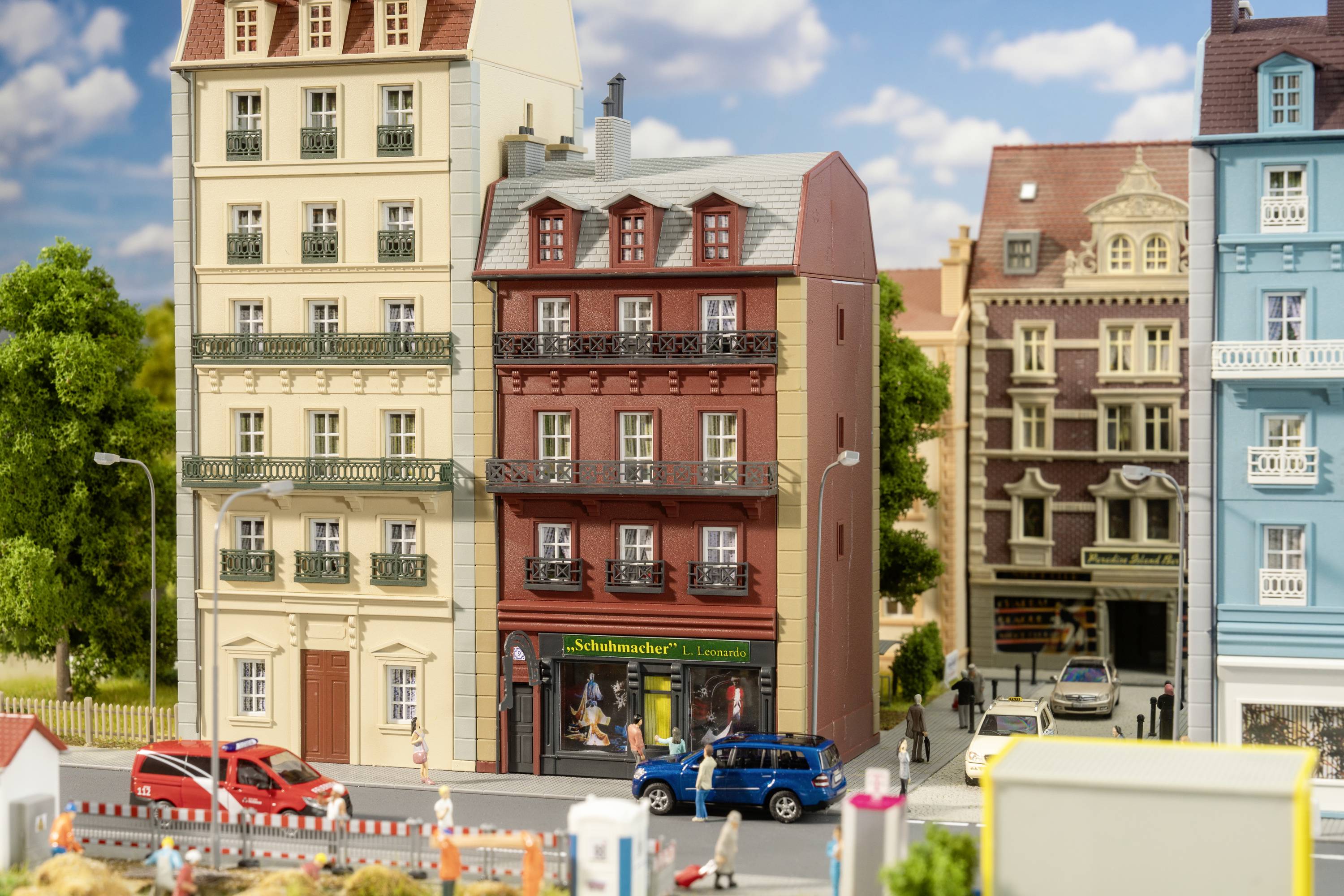 A miniature town displays detailed buildings, cars, and trees. In the foreground, red and blue cars drive near a construction site.