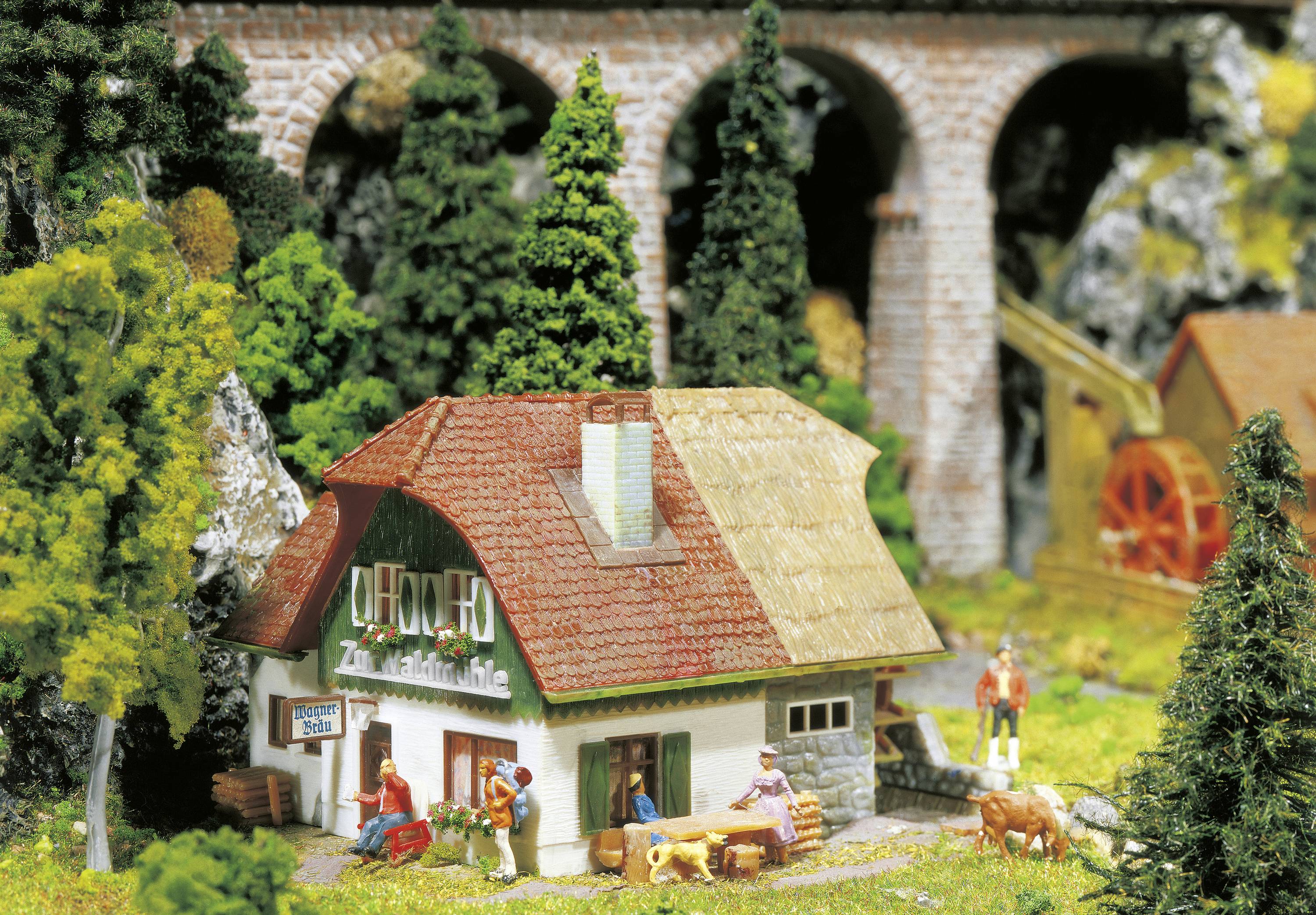 A miniature scene depicts a half-timbered house in a rural setting with pine trees and a viaduct in the background. Figurines bring the scene to life.