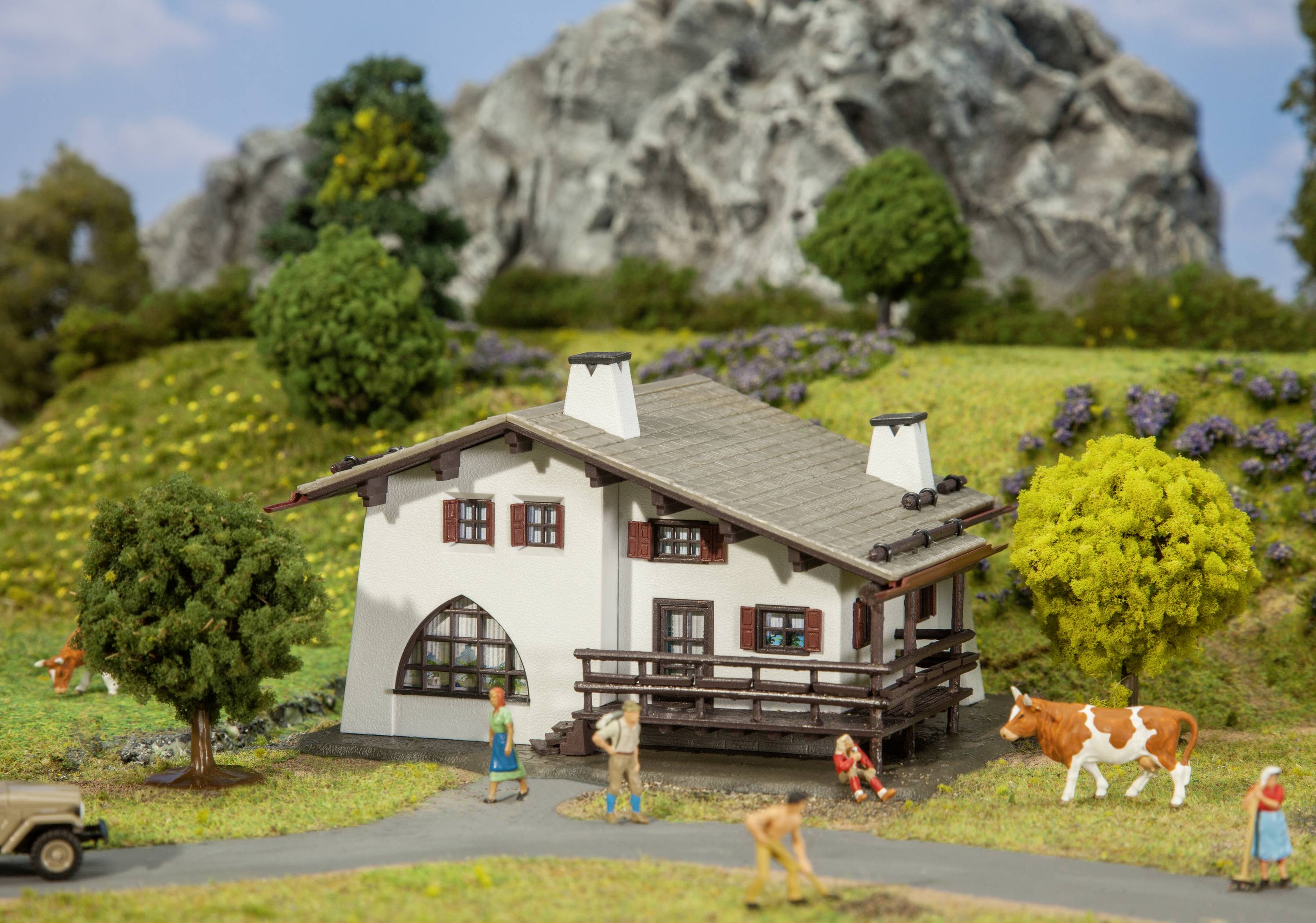 A small white house with a balcony stands in a miniature landscape with trees, flowers, a cow, and miniature figures.