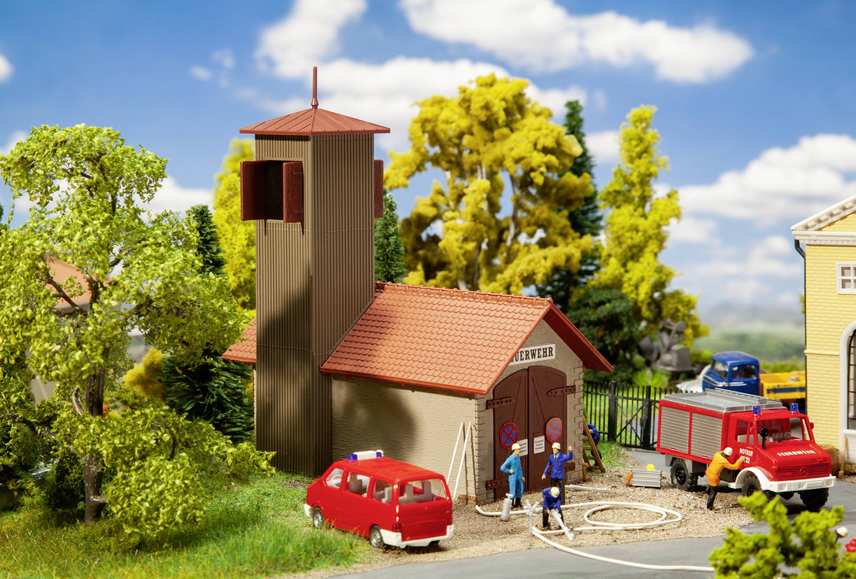 Model of a fire station with two red fire engines in the foreground. Firefighters with hoses are standing nearby.