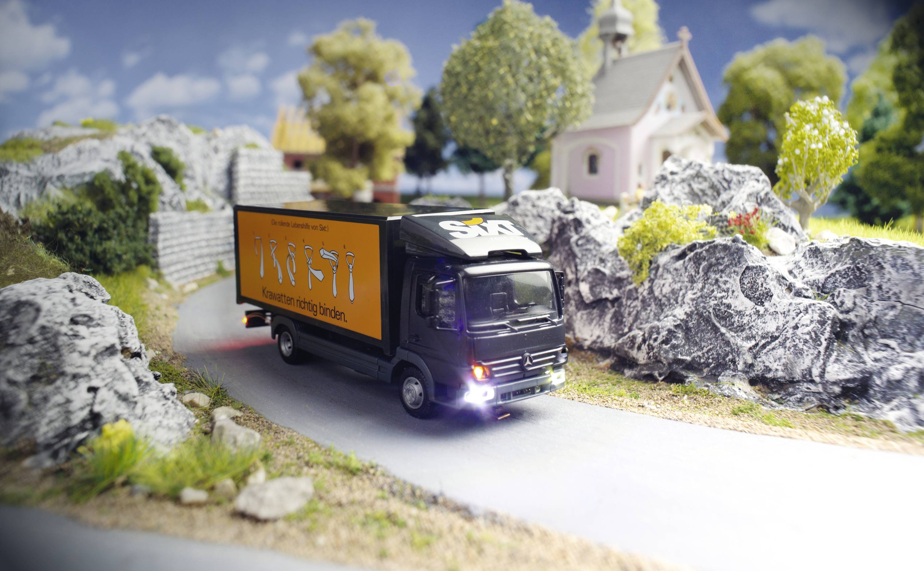 A model lorry is driving along a road through a landscape with rocks, trees, and small buildings in the background.